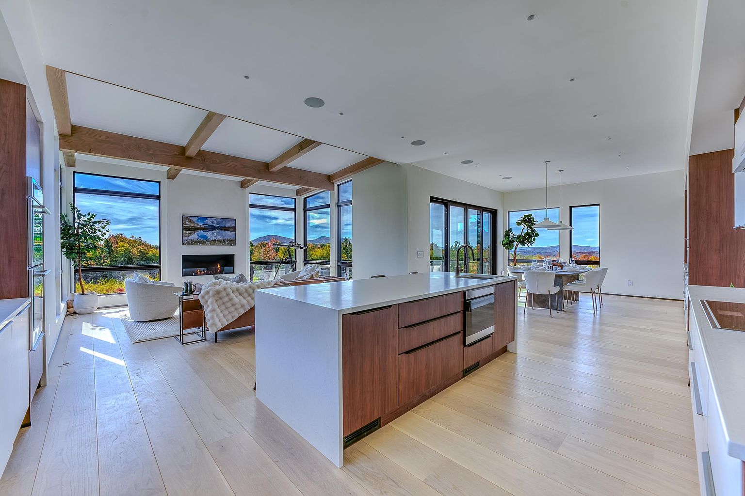 This expansive, open-concept living space seamlessly blends a modern kitchen, dining area, and living room, all bathed in natural light from floor-to-ceiling windows. The kitchen features a large, wood-paneled island with a quartz countertop, while the living area is anchored by a sleek fireplace and exposed wooden ceiling beams. The overall aesthetic is contemporary and airy, offering panoramic views of the surrounding landscape.