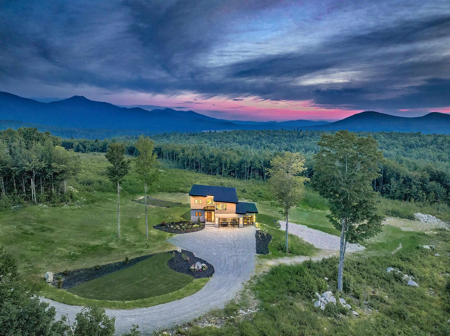 This stunning aerial view captures a modern, multi-level home nestled in a vast, lush landscape against a dramatic sunset backdrop. The property features a clean, contemporary architectural design with a gravel driveway, manicured landscaping, and expansive mountain views. The cinematic perspective highlights the home's secluded and serene setting, emphasizing its integration with the surrounding natural environment.