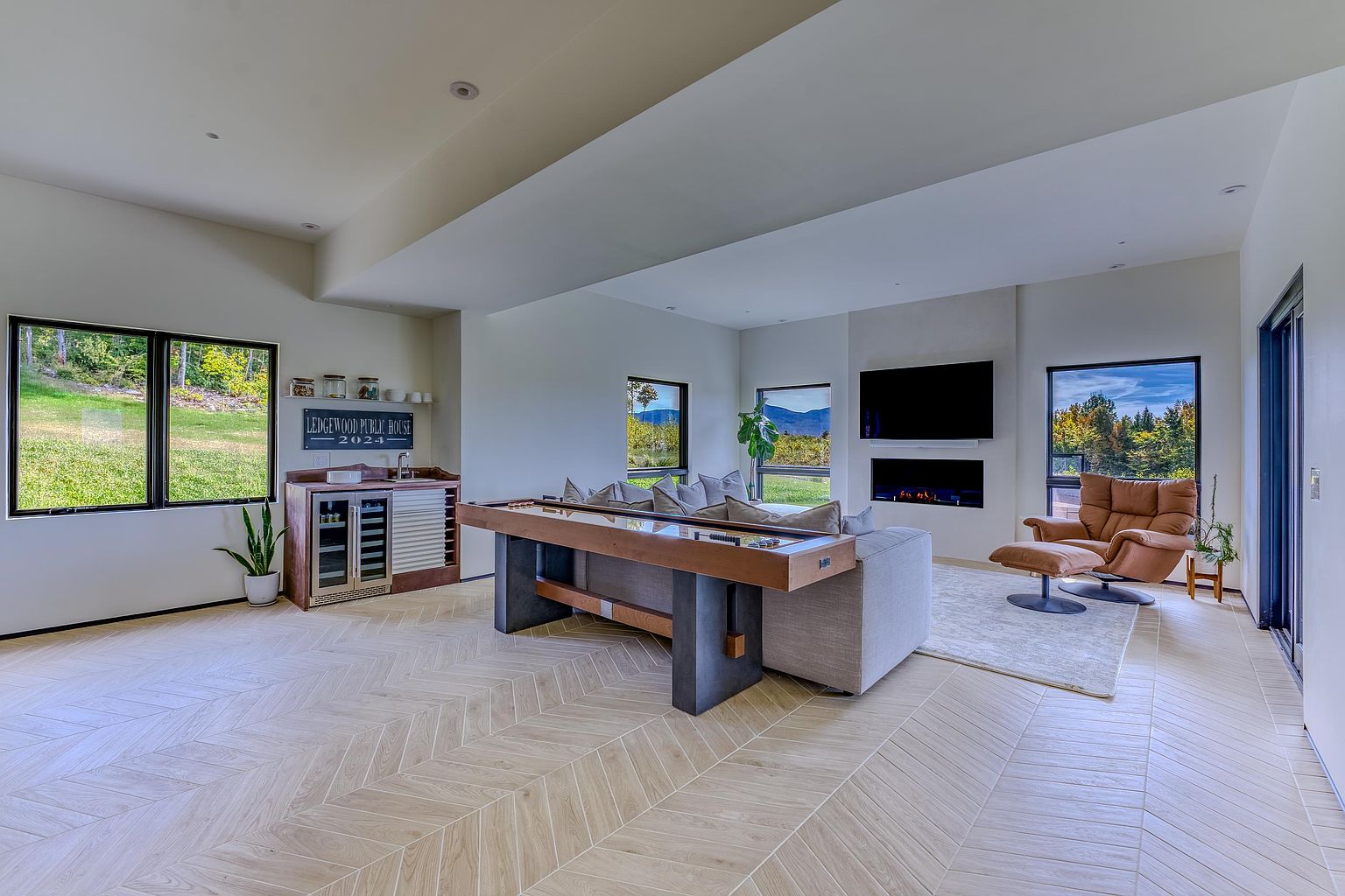 This spacious, modern living room features light-toned herringbone flooring and a large, comfortable sofa positioned behind a shuffleboard table. A sleek fireplace is integrated into the wall beneath a mounted television, while large windows provide expansive views of the surrounding landscape. The room is designed with a contemporary aesthetic, incorporating a small wet bar area and a cozy leather lounge chair, creating an inviting space for relaxation and entertainment.