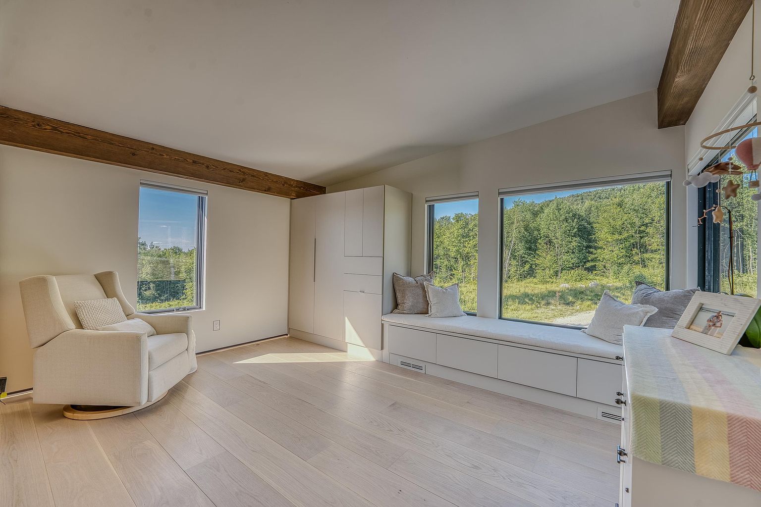 This bright and airy room features a minimalist design with light wood flooring, a comfortable cream-colored armchair, and a built-in window seat with storage underneath. Large windows provide an expansive view of the surrounding forest, while exposed wooden ceiling beams add a touch of rustic warmth to the modern space. The room is bathed in natural light, creating a serene and inviting atmosphere.