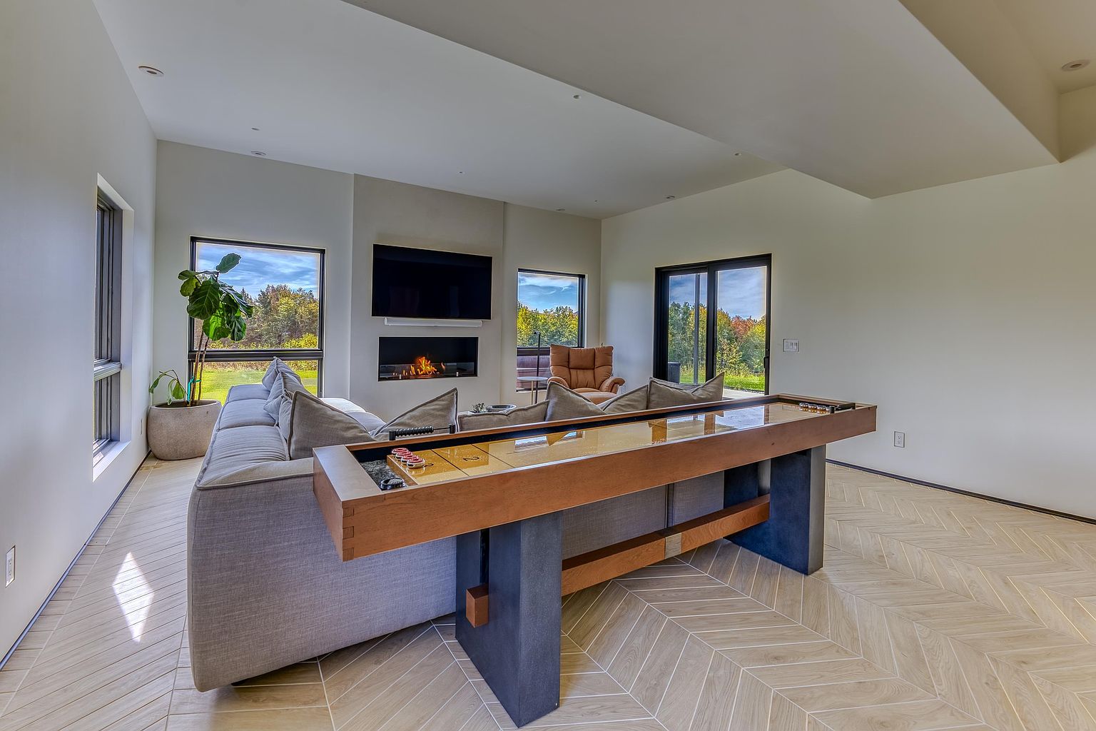 This modern, open-concept living room features a sleek fireplace integrated into a feature wall below a wall-mounted television. A large, comfortable sectional sofa is positioned behind a custom wooden shuffleboard table, creating a unique blend of relaxation and entertainment space. The room is brightened by large windows and glass doors that offer scenic views of the surrounding landscape, all set against light-toned herringbone flooring.