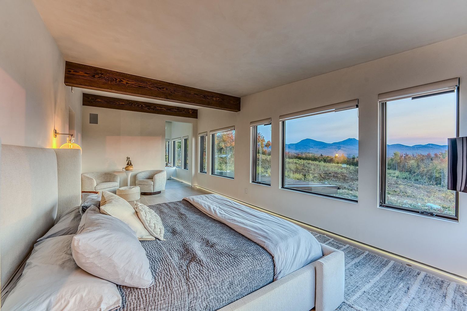 This luxurious primary bedroom features a minimalist, contemporary design with a plush king-sized bed as the focal point. Large, expansive windows offer breathtaking panoramic views of the surrounding mountains and forest, while exposed wooden ceiling beams add a touch of rustic warmth to the clean, modern aesthetic. The room seamlessly transitions into a cozy seating area, creating a serene and sophisticated retreat.