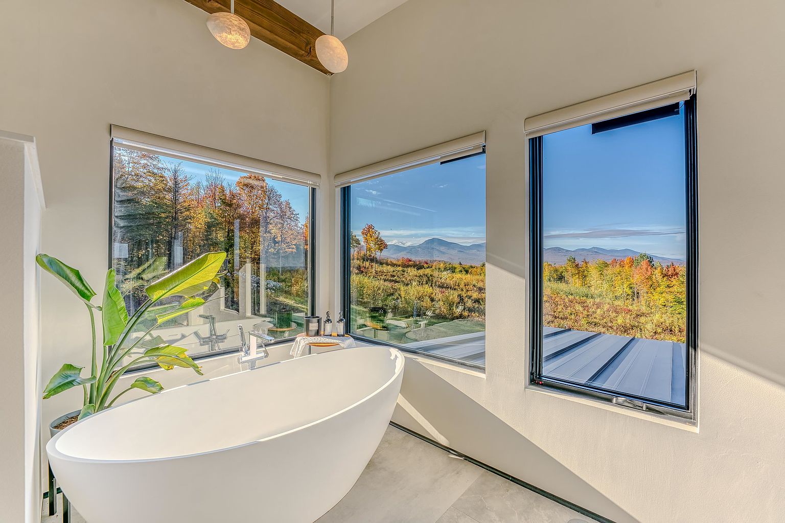This luxurious primary bathroom features a sleek, white freestanding soaking tub positioned perfectly to take advantage of expansive, panoramic mountain views through large, corner-wrapping windows. The minimalist design is complemented by a vibrant potted plant and modern pendant lighting, creating a serene and spa-like atmosphere. The bright, neutral walls and clean lines emphasize the connection between the indoor sanctuary and the natural landscape outside.