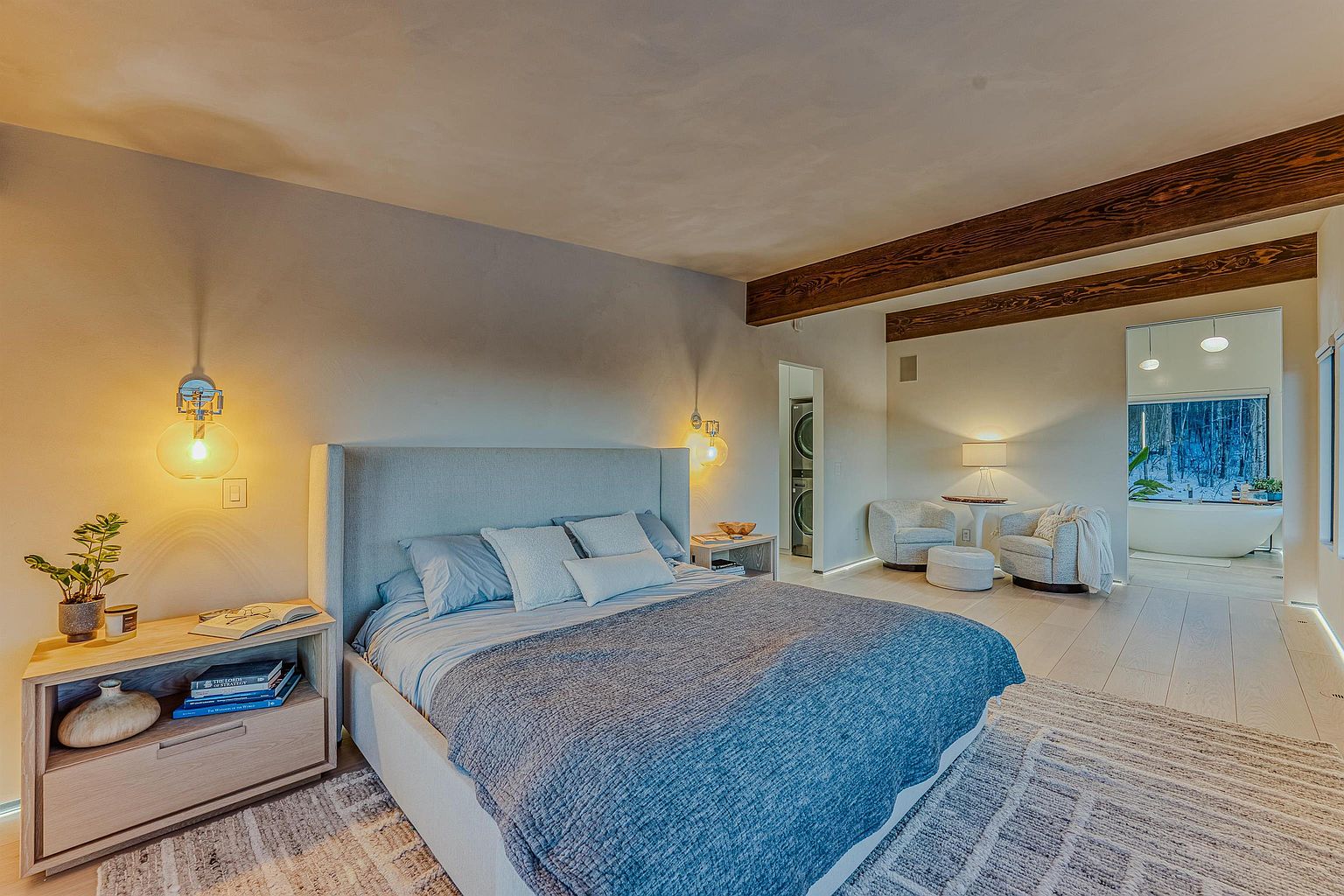 This serene primary bedroom features a contemporary design with a plush upholstered bed, warm wood nightstands, and exposed wooden ceiling beams. The room flows seamlessly into an adjacent seating area and a bright, spa-like bathroom visible through an open doorway. The neutral color palette and soft lighting create a tranquil, high-end atmosphere.