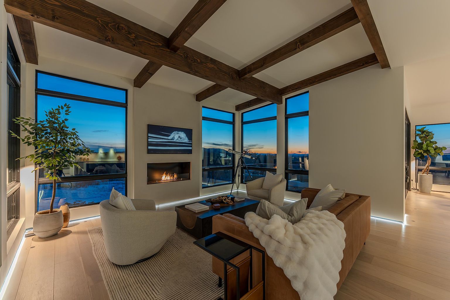 This sophisticated living room features a modern, open-concept design with exposed wooden ceiling beams and expansive floor-to-ceiling windows that frame a stunning twilight view. The space is anchored by a cozy leather sofa with a plush throw, two contemporary armchairs, and a sleek linear fireplace set into the wall. A telescope positioned near the window adds a touch of intellectual charm, while the warm ambient lighting creates an inviting and luxurious atmosphere.