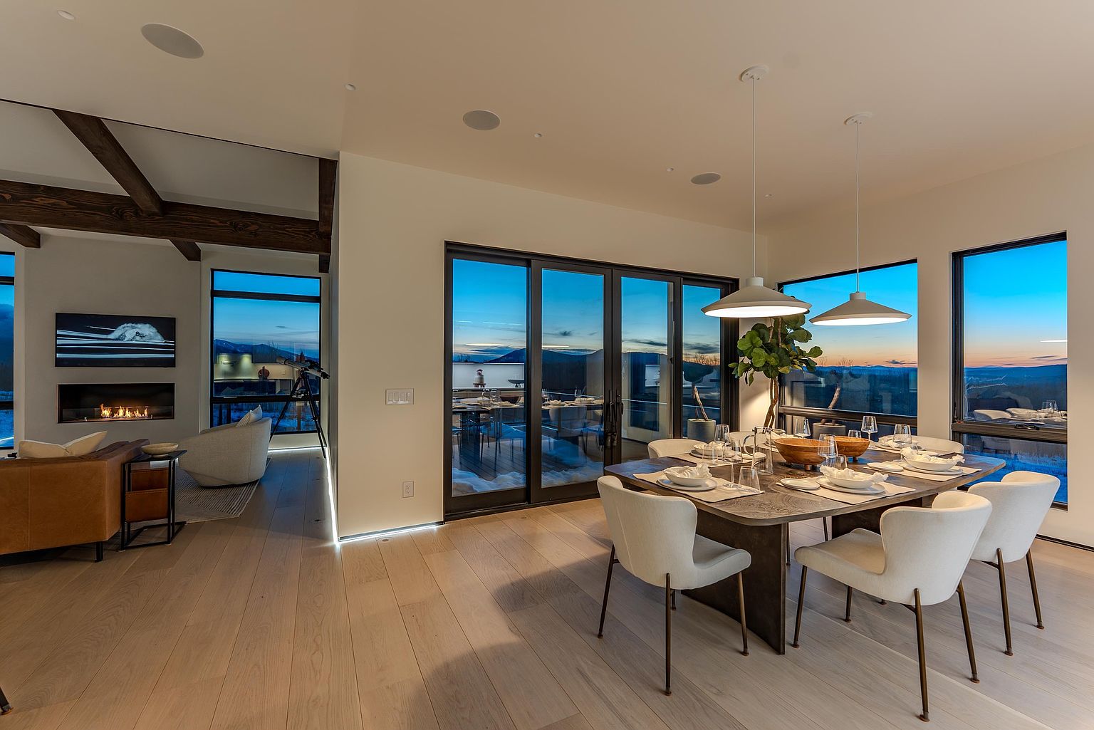 This elegant dining area features a modern wooden table surrounded by chic, cream-colored upholstered chairs, illuminated by minimalist pendant lighting. The open-concept space seamlessly transitions into a cozy living area with a fireplace, while floor-to-ceiling glass doors reveal a stunning sunset view over a mountainous landscape. The interior design emphasizes a sophisticated, contemporary aesthetic with warm wood flooring and clean, neutral tones.