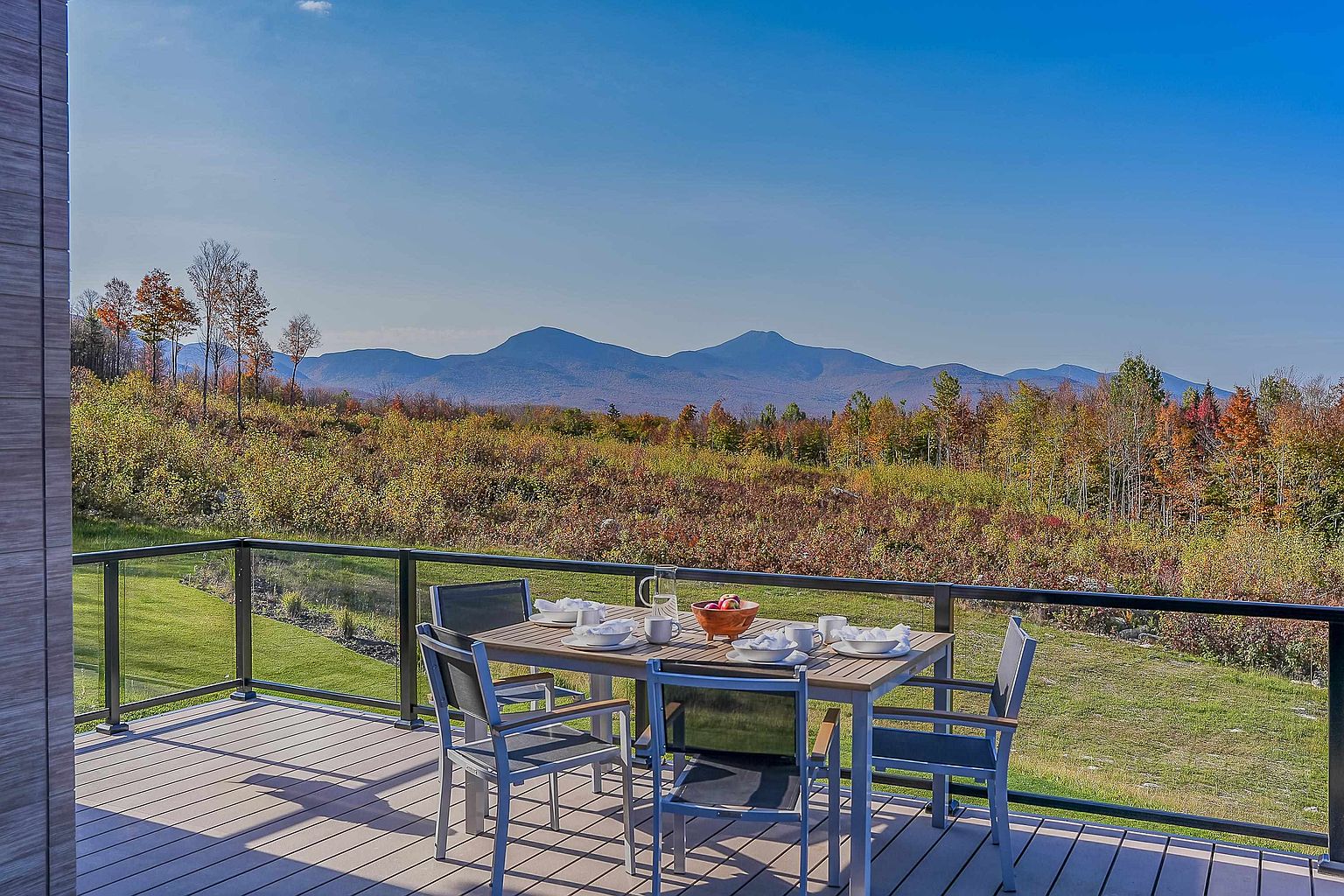 This image showcases a spacious, modern deck featuring a dining set arranged for an outdoor meal, set against a breathtaking panoramic view of rolling mountains and autumn-colored foliage. The deck is enclosed by a sleek glass railing, ensuring an unobstructed connection to the natural landscape. The scene evokes a serene, high-end lifestyle, perfect for enjoying morning coffee or sunset dinners in a tranquil, scenic setting.
