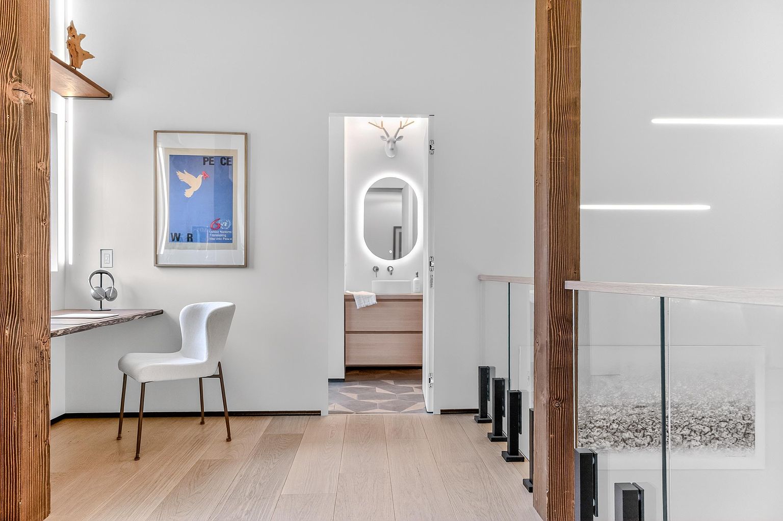 This bright and modern home office space features a minimalist desk with a live-edge wood top and a sleek white chair, set against a clean white wall adorned with a framed peace poster. A doorway leads into a stylish bathroom with a backlit mirror and a unique deer-head wall mount, while the foreground showcases a glass-railed staircase with warm wood accents. The overall aesthetic is contemporary and airy, emphasizing clean lines and natural textures.