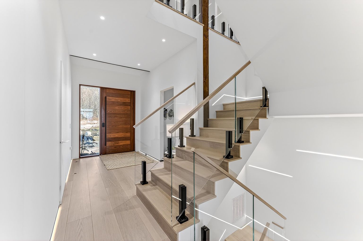 This modern entryway features a striking floating staircase with light wood treads, glass railings, and black metal supports. A large, warm-toned wooden front door with a side window creates a welcoming focal point, while recessed lighting and sleek wall-mounted LED strips provide a clean, contemporary aesthetic. The perspective captures the transition from the front door into the open, airy staircase area, emphasizing the home's high-end architectural design.