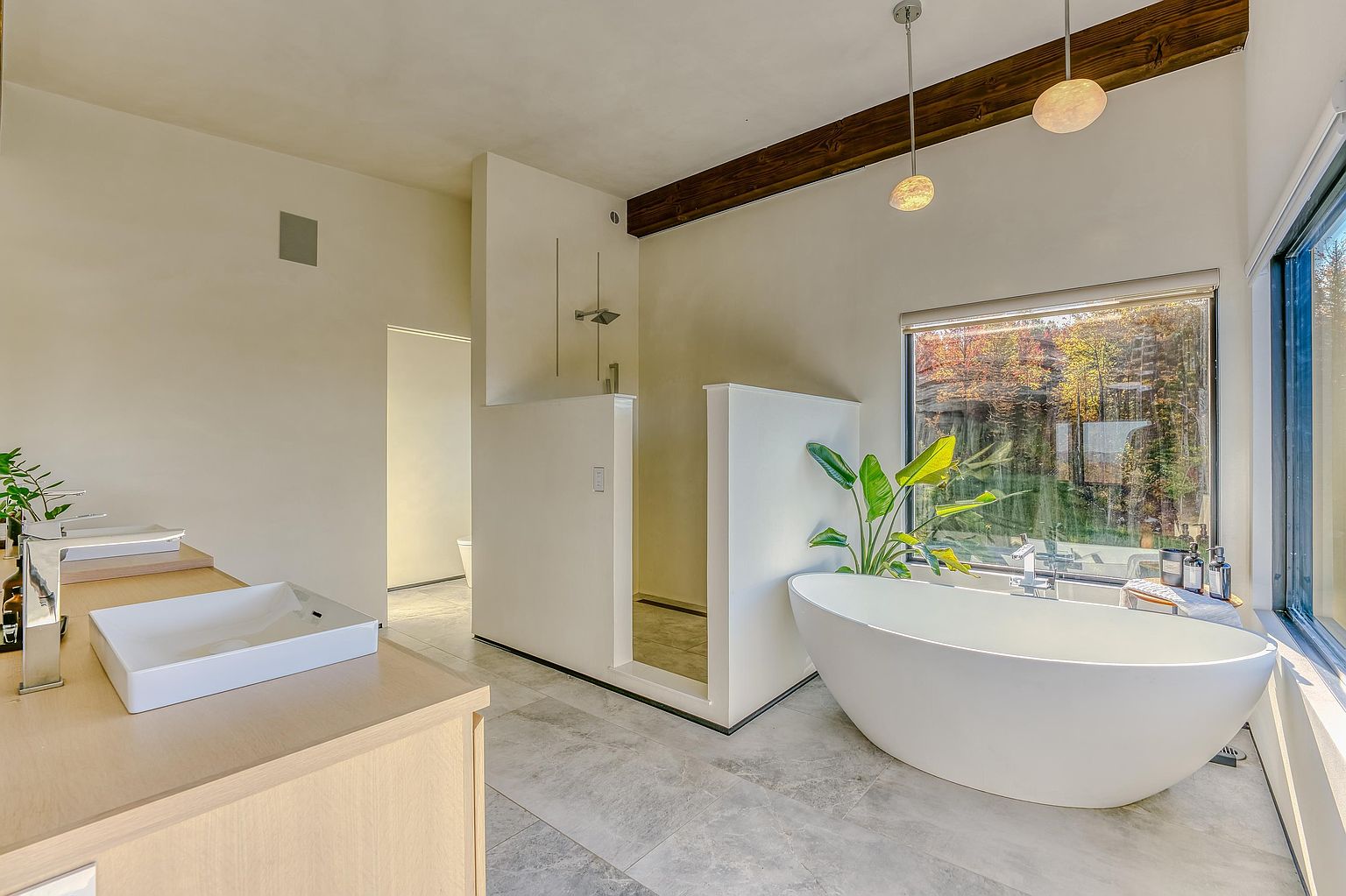 This luxurious primary bathroom features a sleek, modern design with a freestanding soaking tub positioned next to a large window that offers a serene view of the surrounding woods. The space includes a minimalist vanity with a vessel sink and a walk-in shower enclosed by clean, white partition walls. A rustic wooden ceiling beam adds a touch of warmth to the contemporary aesthetic, creating a spa-like atmosphere.