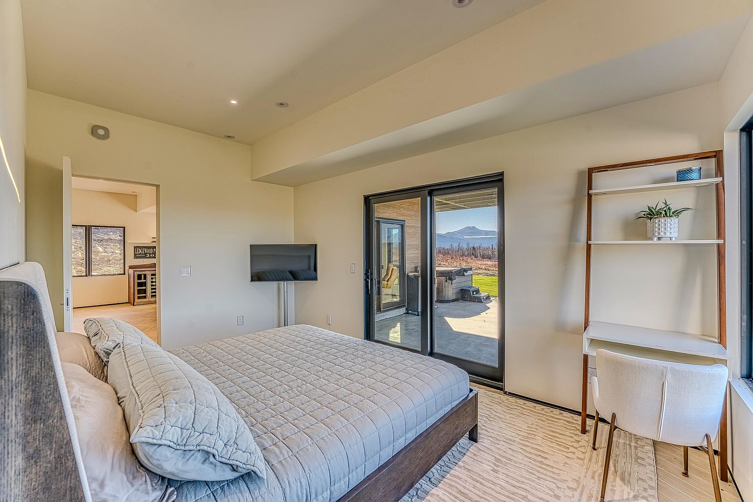 This modern primary bedroom features a neutral color palette, a plush quilted bed, and a sleek desk area with open shelving. Large sliding glass doors provide seamless indoor-outdoor flow to a patio with mountain views, while an adjacent doorway reveals a glimpse of a secondary room. The space is bright and airy, emphasizing a contemporary design with clean lines and functional living areas.