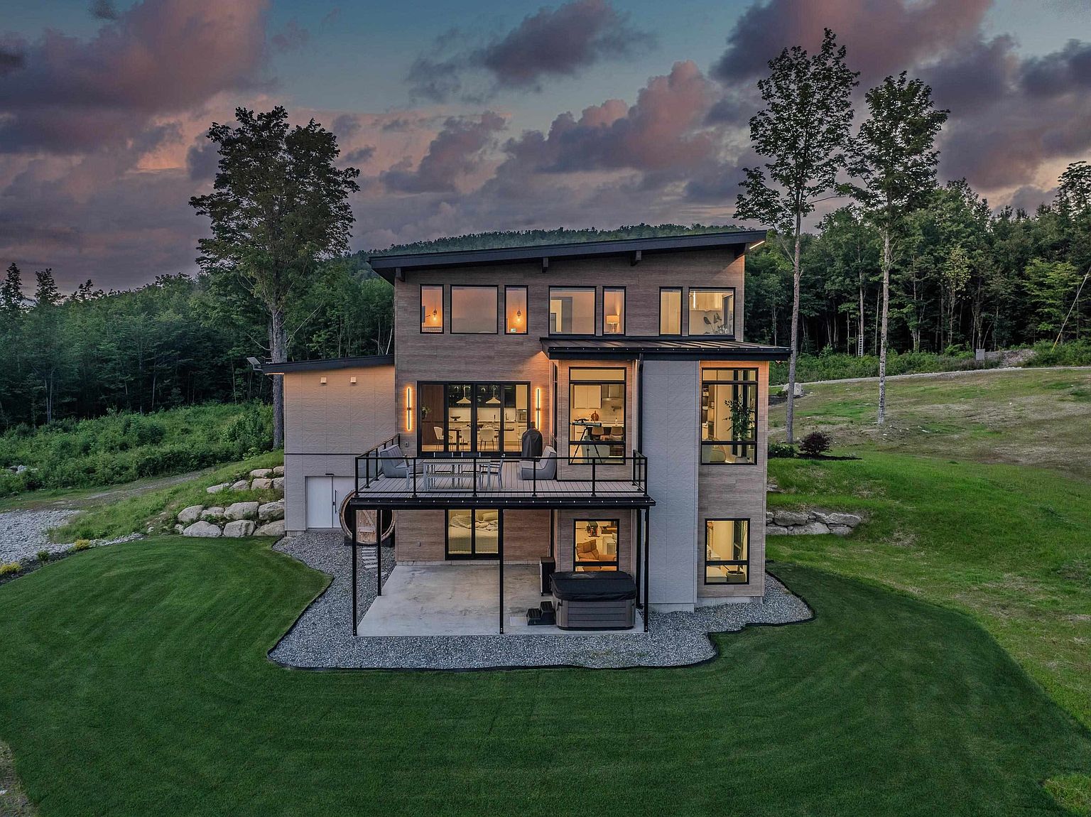 This stunning contemporary home is captured from an elevated rear perspective at dusk, showcasing its modern architectural design with clean lines and a mix of wood and stone textures. The property features a spacious elevated deck with a black railing, a lower-level concrete patio housing a hot tub, and large floor-to-ceiling windows that glow warmly against the twilight sky. Set against a backdrop of lush forest and rolling green lawns, the home exudes a sophisticated, tranquil, and luxurious atmosphere.