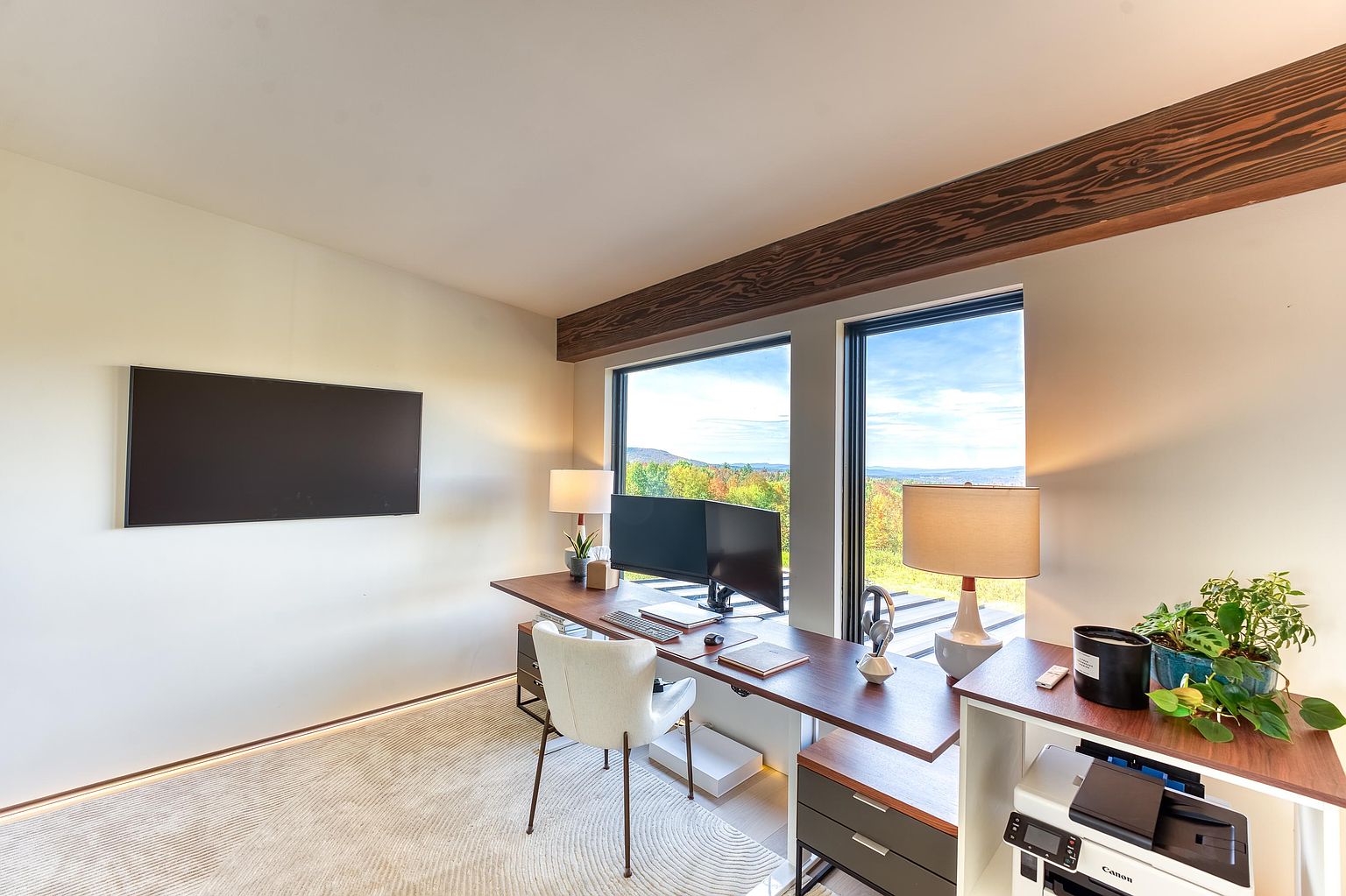 This modern home office features a sleek, long wooden desk positioned against large windows that offer a stunning, elevated view of the surrounding landscape. The room is accented by a rustic wooden ceiling beam, a minimalist white chair, and a wall-mounted television, creating a professional yet serene workspace. The clean lines and neutral color palette emphasize a contemporary and productive atmosphere.
