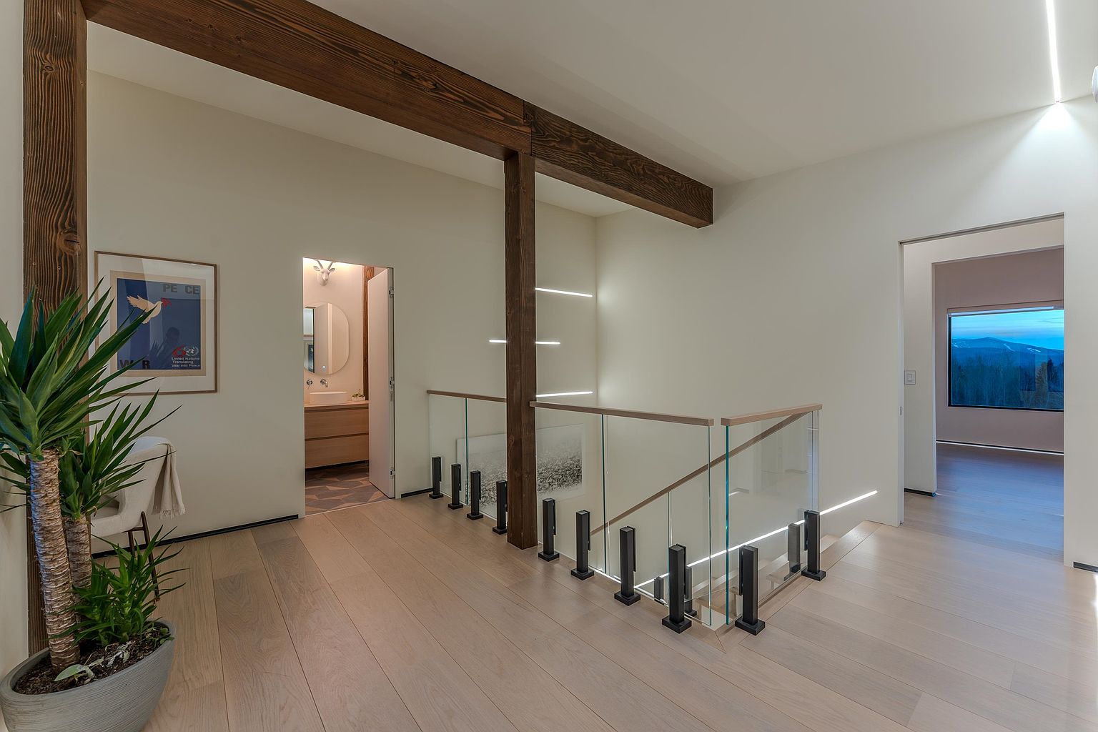 This modern, minimalist hallway features light-toned hardwood flooring and a striking staircase with a glass railing and dark metal posts. A prominent exposed wooden beam adds architectural character, while a doorway leads to a bright bathroom and another opens to a room with a scenic mountain view. The space is clean, airy, and sophisticated, emphasizing high-end design and natural light.