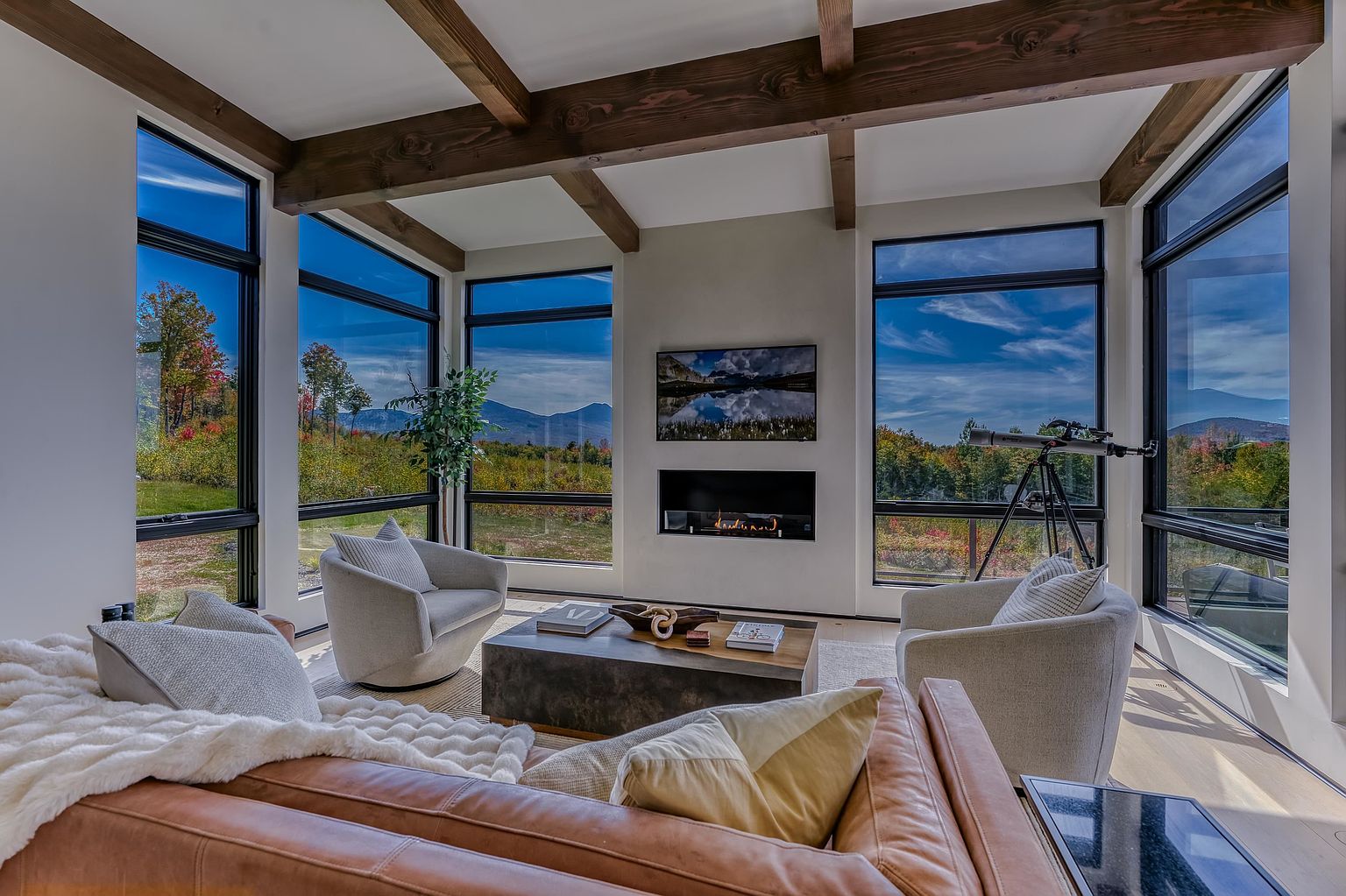 This sophisticated living room features a modern, open-concept design with floor-to-ceiling windows that frame breathtaking mountain and forest views. The space is anchored by a sleek, linear fireplace and a wall-mounted television, complemented by warm wood ceiling beams and a plush leather sofa. The combination of contemporary furnishings and natural, panoramic scenery creates an inviting and luxurious atmosphere perfect for relaxation.
