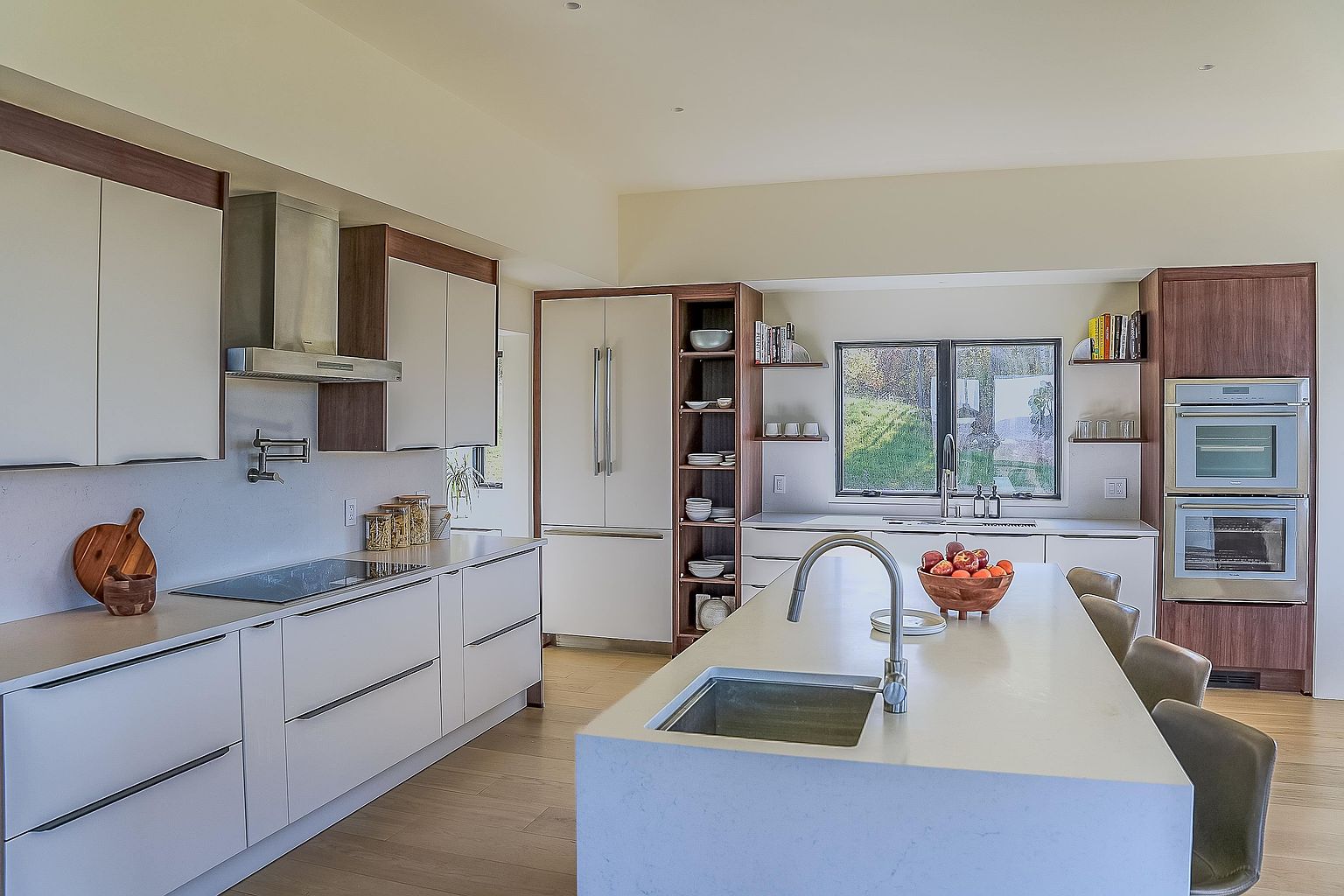 This modern, high-end kitchen features a sleek, minimalist design with a large central island topped with a light-colored quartz countertop and a built-in sink. The space is accented by warm wood cabinetry, stainless steel appliances, and open shelving, creating a sophisticated and functional atmosphere. A large window above the sink provides ample natural light, while the open-concept layout makes it ideal for both cooking and entertaining.
