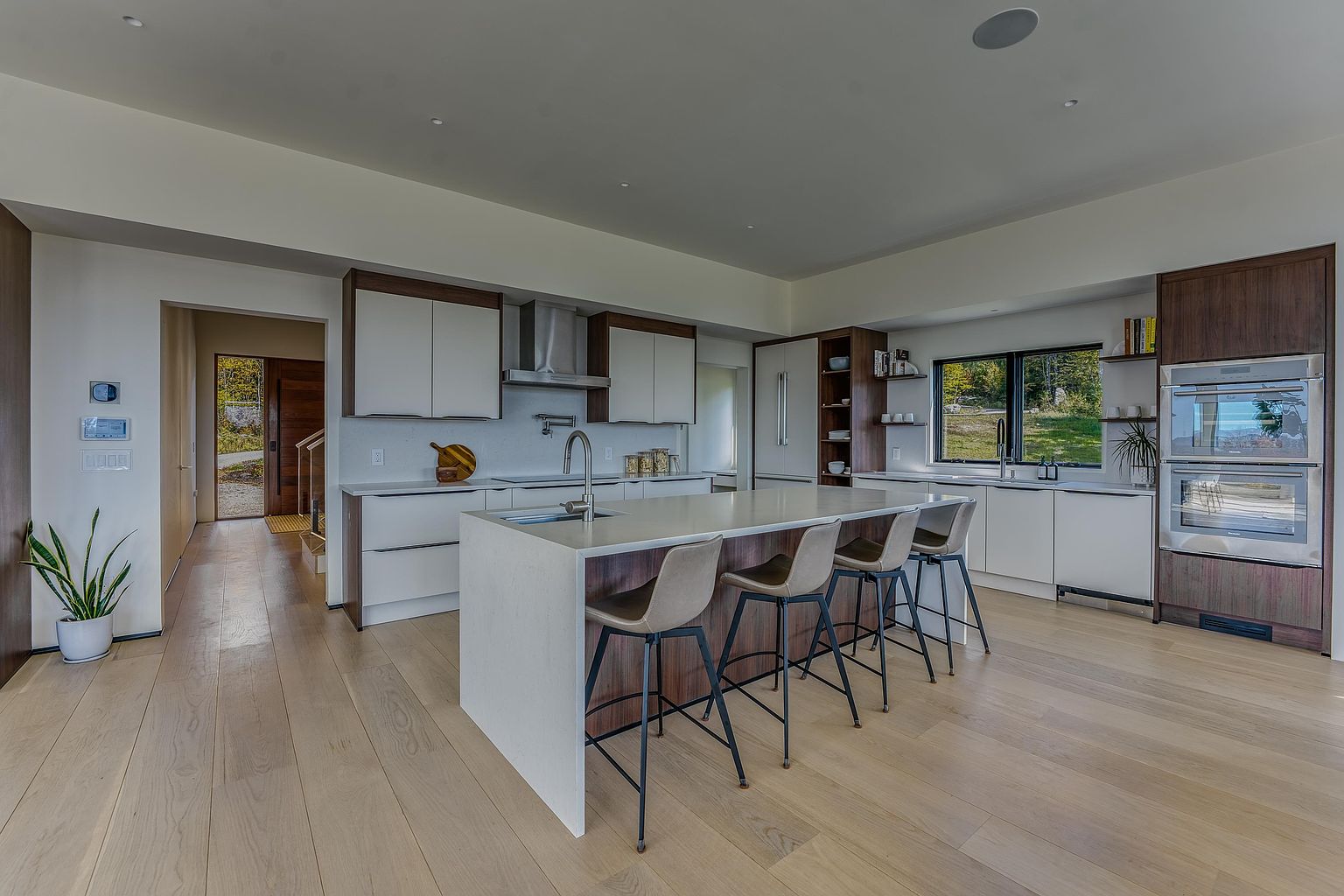 This modern, open-concept kitchen features a sleek island with bar seating, high-end stainless steel appliances, and a sophisticated blend of white and dark wood cabinetry. The space is illuminated by recessed lighting and natural light from a large window, creating a bright and airy atmosphere that flows seamlessly into the adjacent hallway. The minimalist design, light hardwood flooring, and clean lines offer a contemporary and luxurious feel perfect for modern living.
