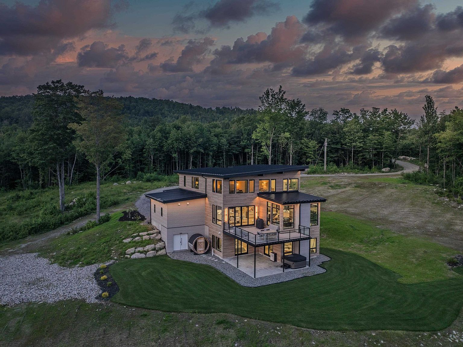 This stunning aerial shot captures a modern, multi-level home nestled in a lush, forested landscape during a dramatic sunset. The property features a contemporary design with clean lines, a spacious elevated deck, and a ground-level patio complete with a hot tub and a barrel sauna. The expansive lawn and surrounding woodland create a serene, private atmosphere, highlighting the home's integration with its natural environment.