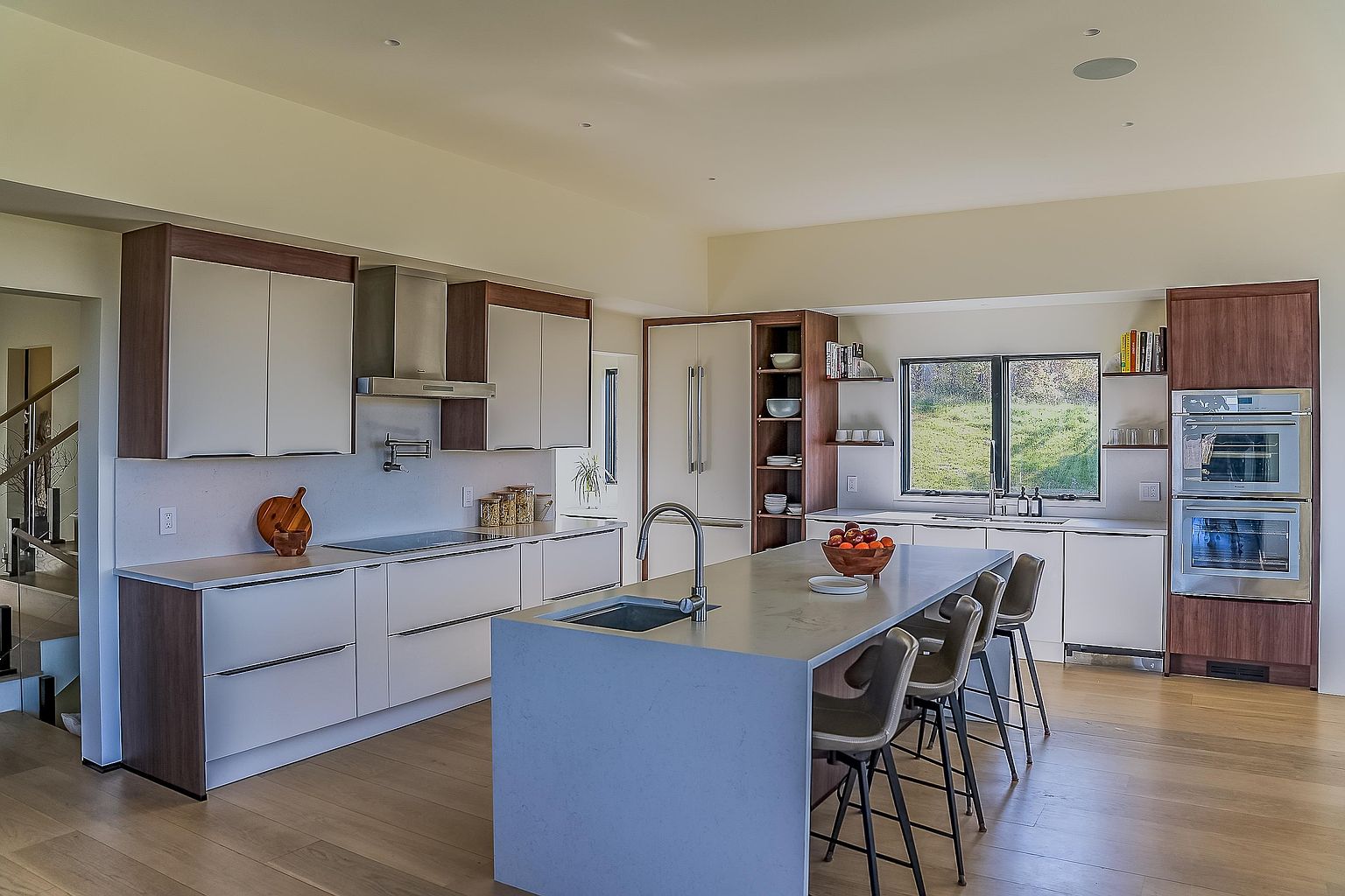 This modern, open-concept kitchen features a sleek design with a large central island, light-toned cabinetry, and warm wood flooring. The space is equipped with high-end stainless steel appliances, including a double wall oven and a professional-grade range hood, while a large window provides natural light and a view of the outdoors. The minimalist aesthetic is complemented by bar seating at the island, creating a functional and inviting atmosphere for cooking and entertaining.