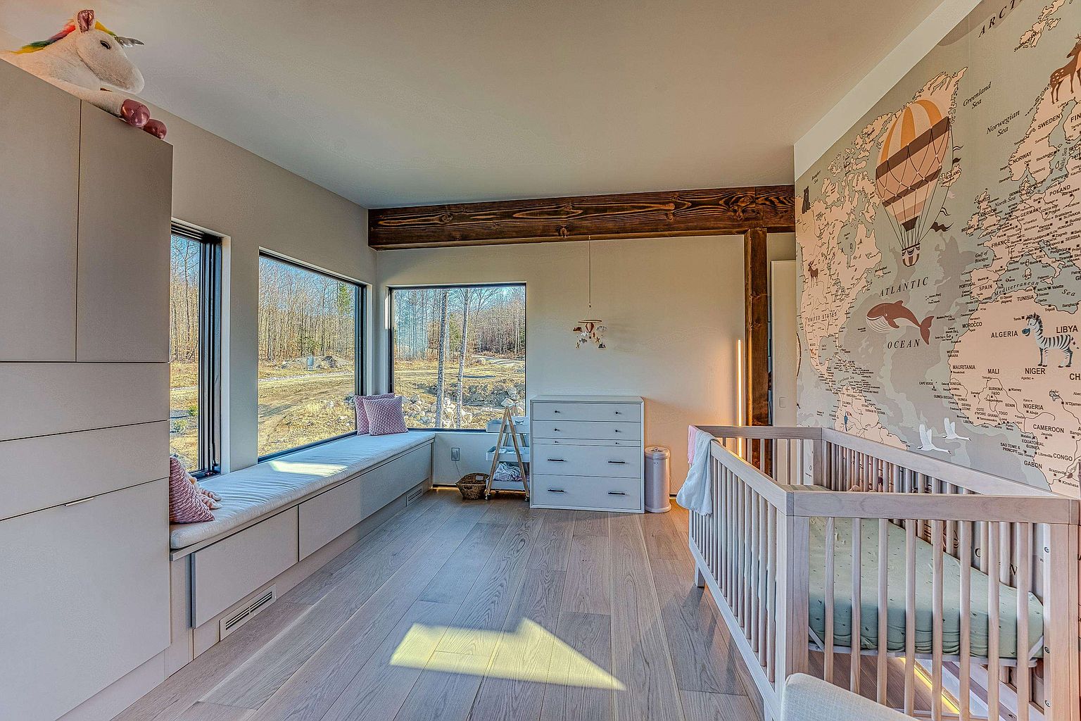 This bright and airy nursery features a large, custom-built window seat that offers expansive views of the surrounding wooded landscape. The room is accented by a charming world map wallpaper, a light-toned wooden crib, and a minimalist white dresser, creating a serene and playful atmosphere. The combination of natural wood flooring, exposed ceiling beams, and abundant natural light gives the space a modern, Scandinavian-inspired aesthetic.