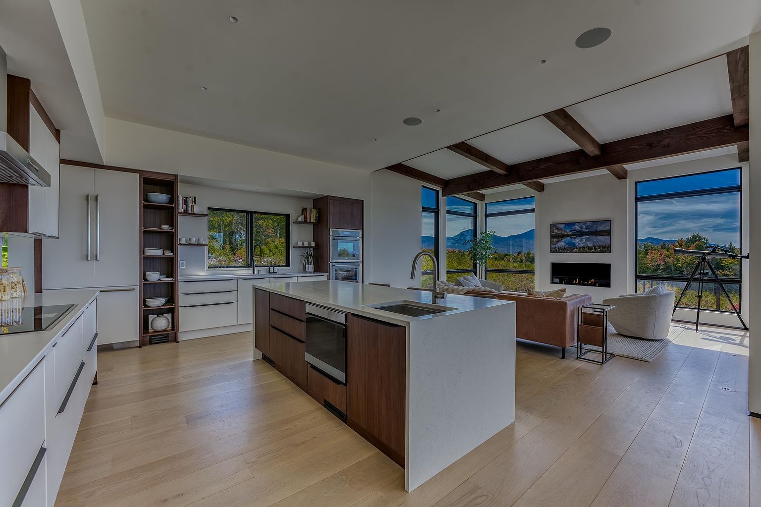 This modern, open-concept kitchen features a large central island with a waterfall quartz countertop and dark wood cabinetry, seamlessly flowing into a bright living area. The space is defined by high ceilings with exposed wooden beams, light hardwood flooring, and expansive floor-to-ceiling windows that frame a stunning mountain view. The design aesthetic is contemporary and airy, emphasizing a sophisticated blend of natural textures and clean, minimalist lines.