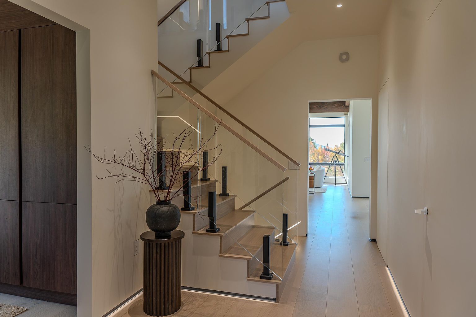 This elegant interior space features a modern staircase with light wood treads, glass railings, and sleek black metal balusters. A decorative vase with branches sits on a fluted pedestal in the foreground, while a bright, open hallway leads toward a sunlit room with a telescope in the distance. The minimalist design, warm wood flooring, and clean lines create a sophisticated and airy atmosphere.