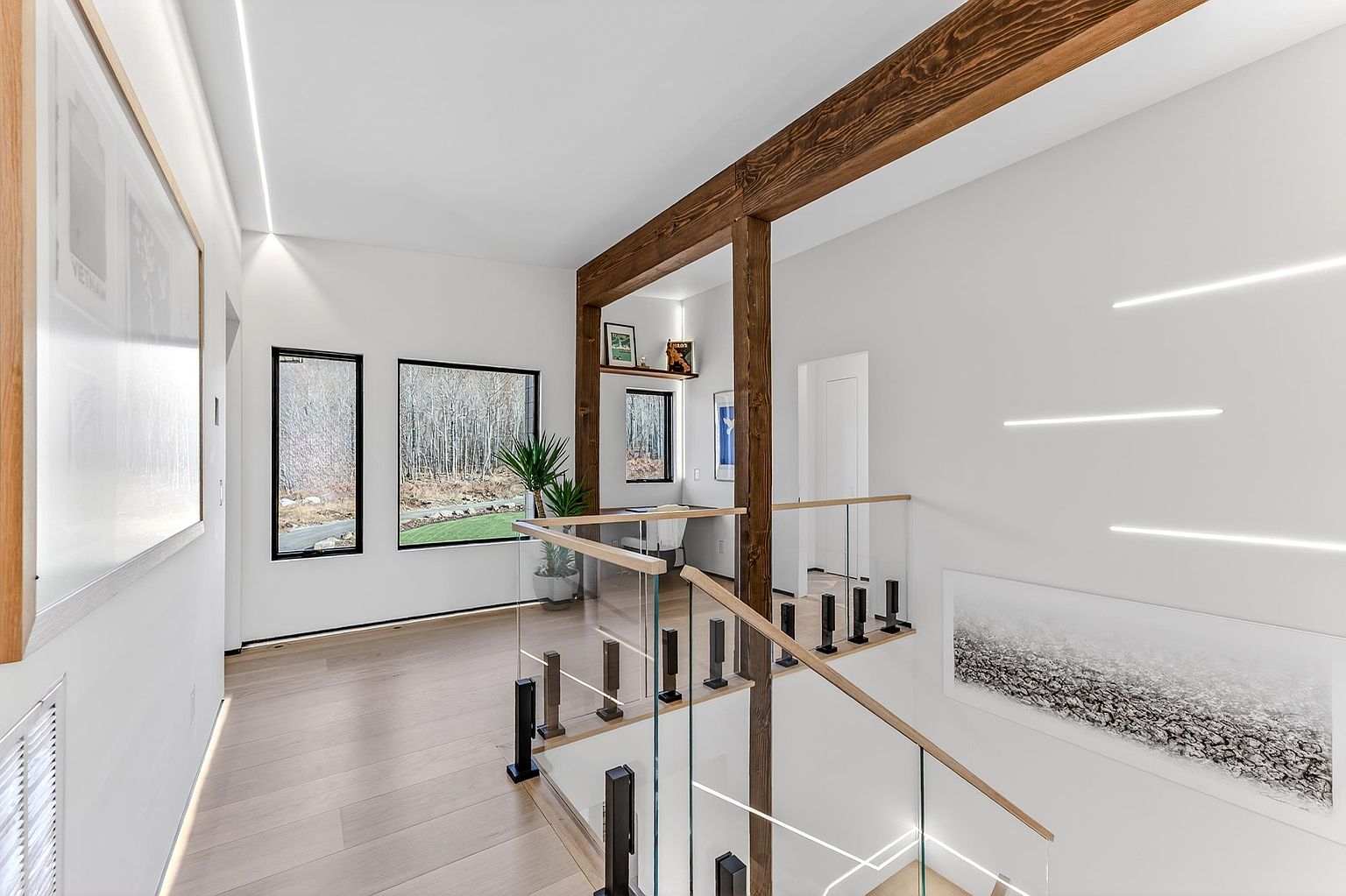 This modern, minimalist hallway features a striking glass-railed staircase and a warm wood-beamed ceiling that adds architectural character. The space is illuminated by sleek, recessed linear lighting and large windows that frame a serene outdoor view, creating an airy and sophisticated atmosphere. A small, integrated workspace nook is tucked neatly into the corner, highlighting the home's efficient and contemporary design.