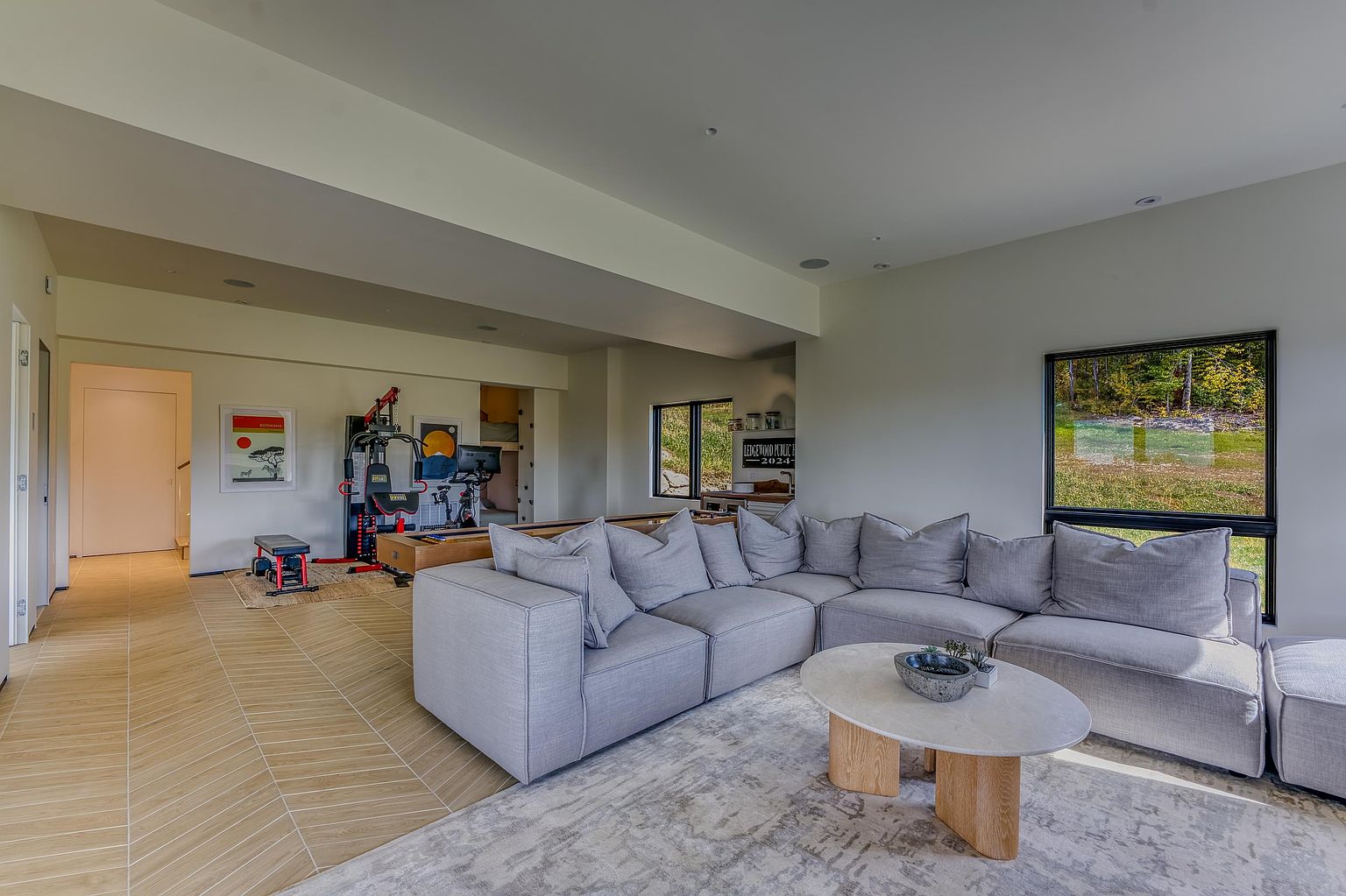 This spacious, open-concept living area features a large, light-gray sectional sofa arranged around a modern oval coffee table on a textured area rug. The room seamlessly transitions into a multi-purpose space that includes a home gym area with exercise equipment and a pool table, all set against a backdrop of light wood-look flooring and neutral walls. Large windows provide ample natural light and views of the surrounding landscape, creating a bright and versatile living environment.