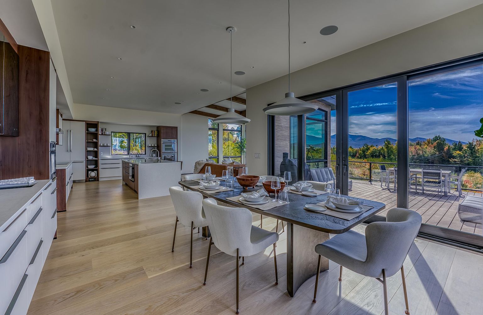 This open-concept space seamlessly blends a modern dining area with a sleek, high-end kitchen, featuring light hardwood floors and minimalist cabinetry. A large, dark-toned dining table serves as the centerpiece, illuminated by contemporary pendant lights, while expansive floor-to-ceiling glass sliding doors offer a breathtaking panoramic view of the mountains and deck. The overall aesthetic is sophisticated and airy, emphasizing a luxurious indoor-outdoor living experience.