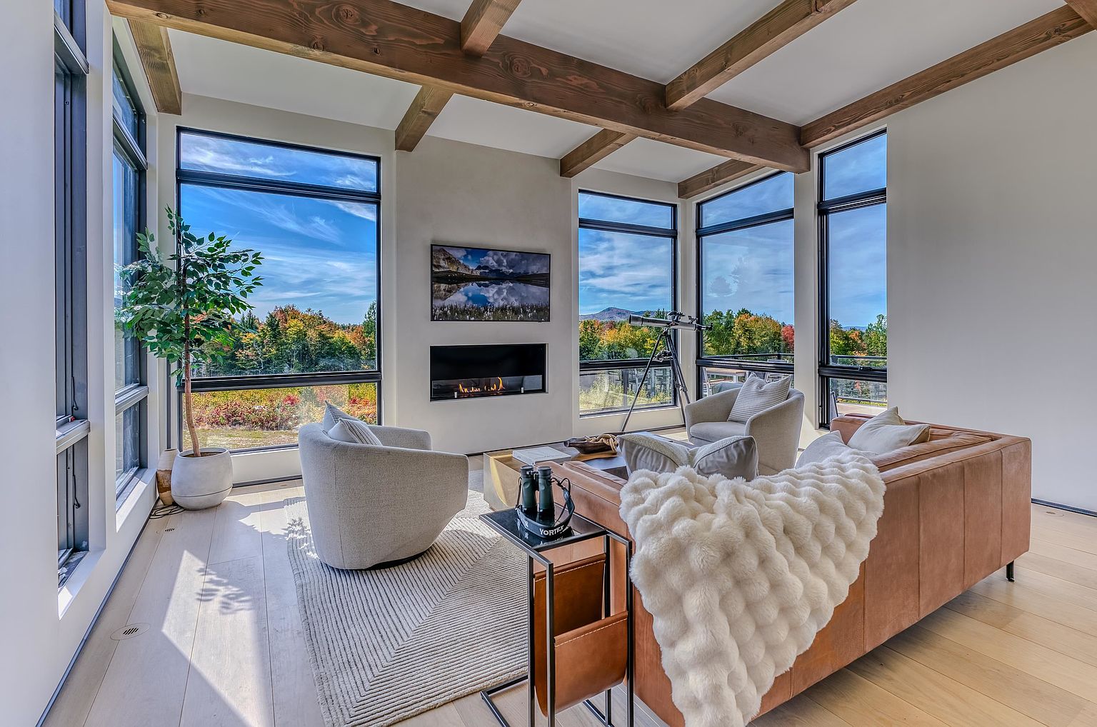 This modern, sun-drenched living room features floor-to-ceiling windows that frame a breathtaking panoramic view of a lush, forested landscape. The space is anchored by a sleek, linear fireplace and exposed wooden ceiling beams, creating a warm, sophisticated atmosphere. A plush leather sofa, textured area rug, and comfortable armchairs provide a cozy seating arrangement perfect for taking in the scenery.