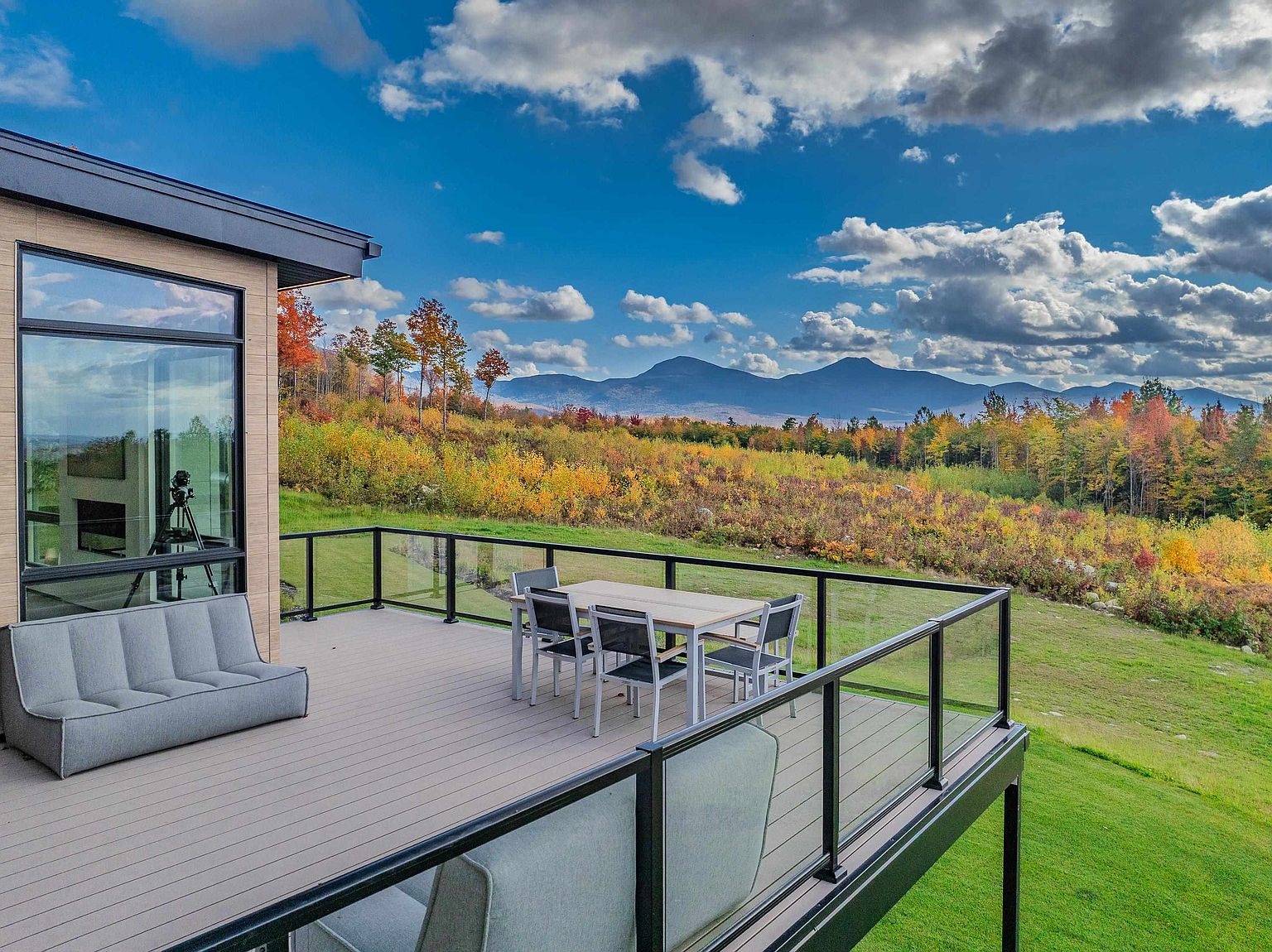 This elevated deck offers a stunning panoramic view of a vibrant, autumn-colored landscape and distant mountain ranges. The space is furnished with a modern outdoor dining set and a sleek, contemporary sofa, all set against a backdrop of expansive glass windows that reflect the scenic beauty. The clean lines of the deck's glass railing and the minimalist architecture create a sophisticated and serene outdoor living experience.