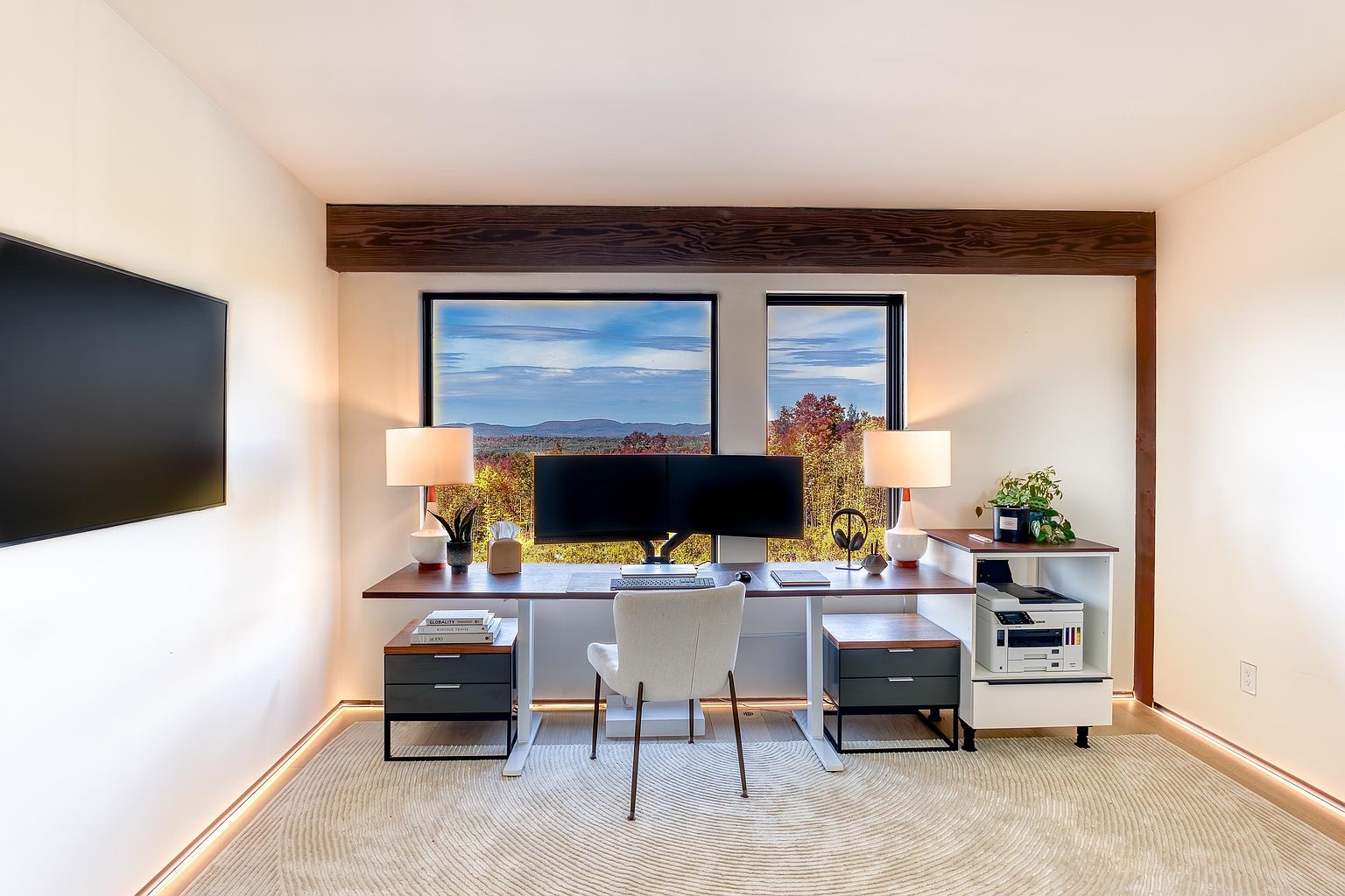 This modern home office features a sleek, minimalist desk setup positioned directly in front of large windows that offer a stunning, panoramic view of a forested mountain landscape. The room is accented by a warm wooden ceiling beam, ambient floor lighting, and a neutral color palette, creating a serene and productive atmosphere. The symmetrical arrangement of lamps and dual monitors emphasizes a clean, professional aesthetic perfect for remote work.