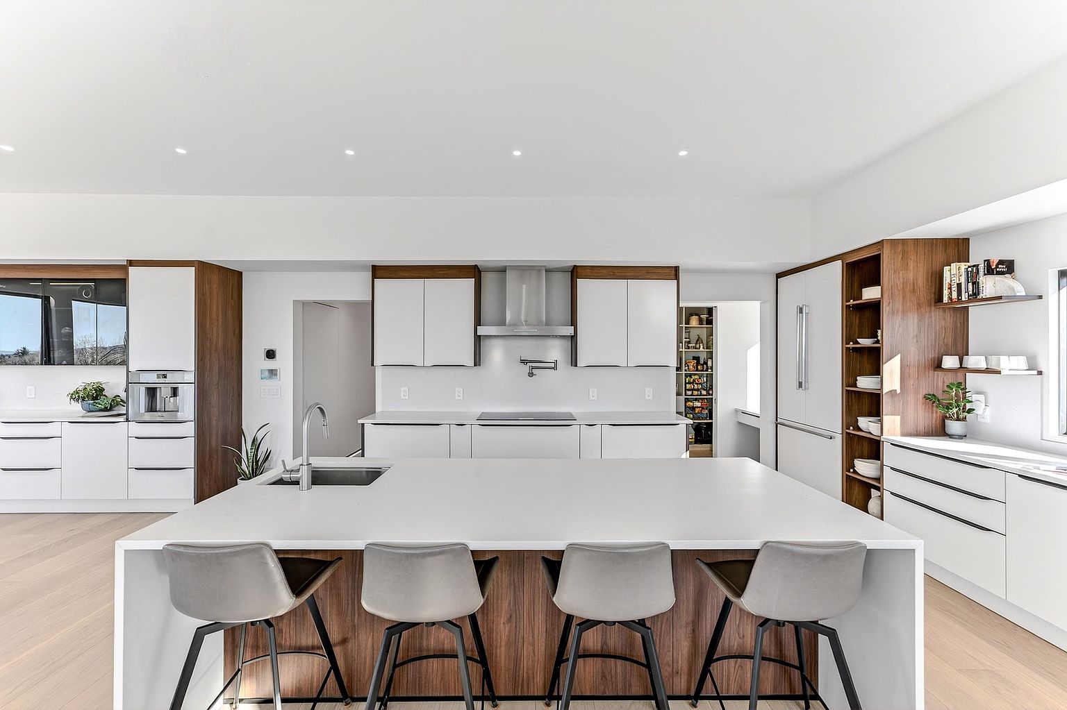 This sleek, modern kitchen features a minimalist design with a large central island, white cabinetry, and warm wood accents. The space is equipped with high-end stainless steel appliances, including a built-in coffee machine and a professional-style range hood, all set against a bright, clean backdrop. The wide-angle perspective highlights the open-concept layout, making it an ideal space for both cooking and entertaining.