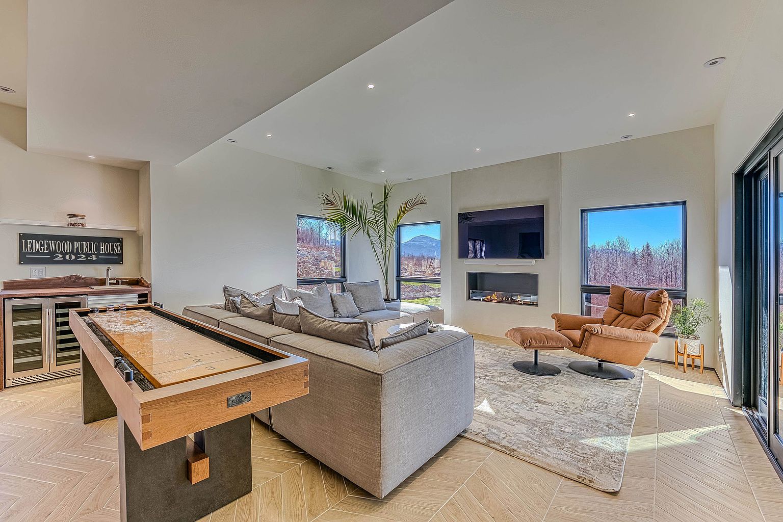 This spacious, modern living room features a large sectional sofa, a comfortable leather lounge chair with an ottoman, and a shuffleboard table, creating an inviting entertainment space. The room is anchored by a sleek fireplace and a wall-mounted television, with expansive windows that frame scenic mountain views. The light-toned herringbone flooring and neutral color palette contribute to a bright, contemporary, and luxurious atmosphere.
