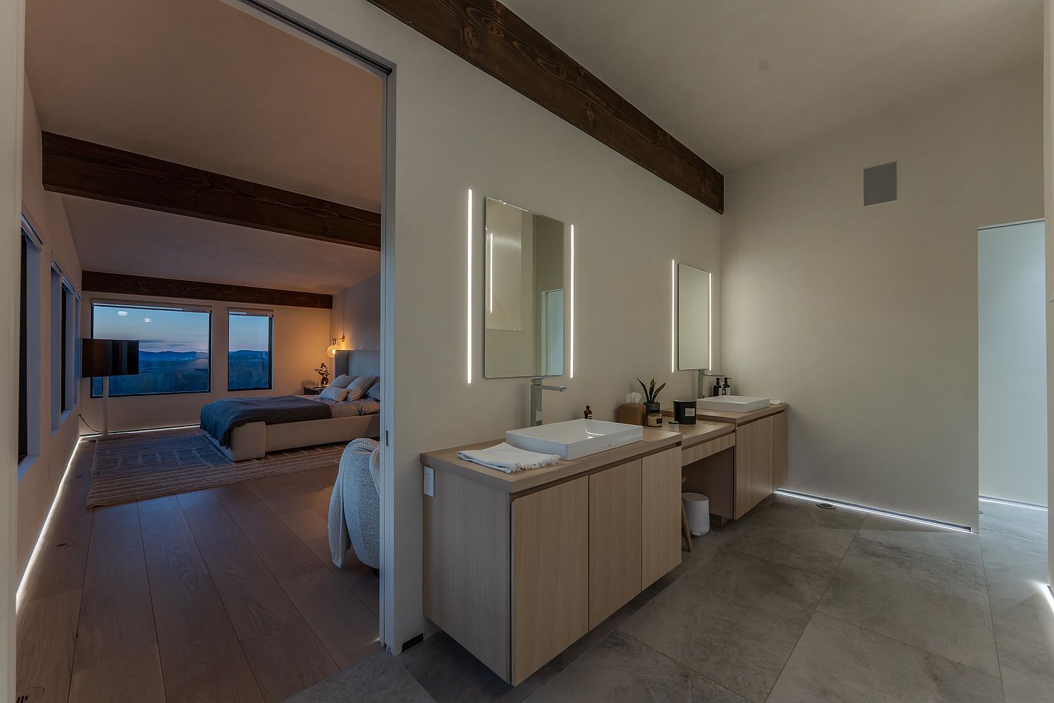 This modern primary bathroom features a sleek, minimalist design with light wood cabinetry, dual vessel sinks, and integrated vertical LED mirror lighting. The space flows seamlessly into an adjacent bedroom, creating an open-concept suite highlighted by warm wood ceiling beams and large-format floor tiles. The perspective captures the transition between the private sleeping area and the spa-like bathroom, emphasizing a sophisticated, high-end aesthetic.