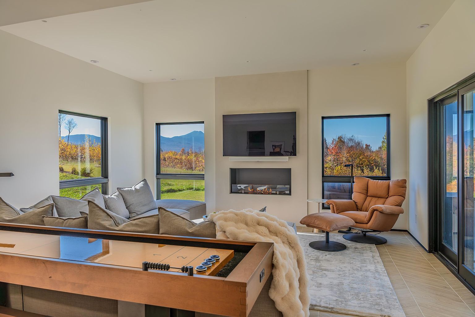 This modern living room features a sleek, minimalist design with a large sectional sofa, a cozy leather lounge chair with an ottoman, and a built-in fireplace beneath a wall-mounted television. A shuffleboard table in the foreground adds a playful element, while expansive windows frame stunning mountain views and vibrant autumn foliage. The space is bathed in natural light, creating an inviting and sophisticated atmosphere perfect for relaxation.