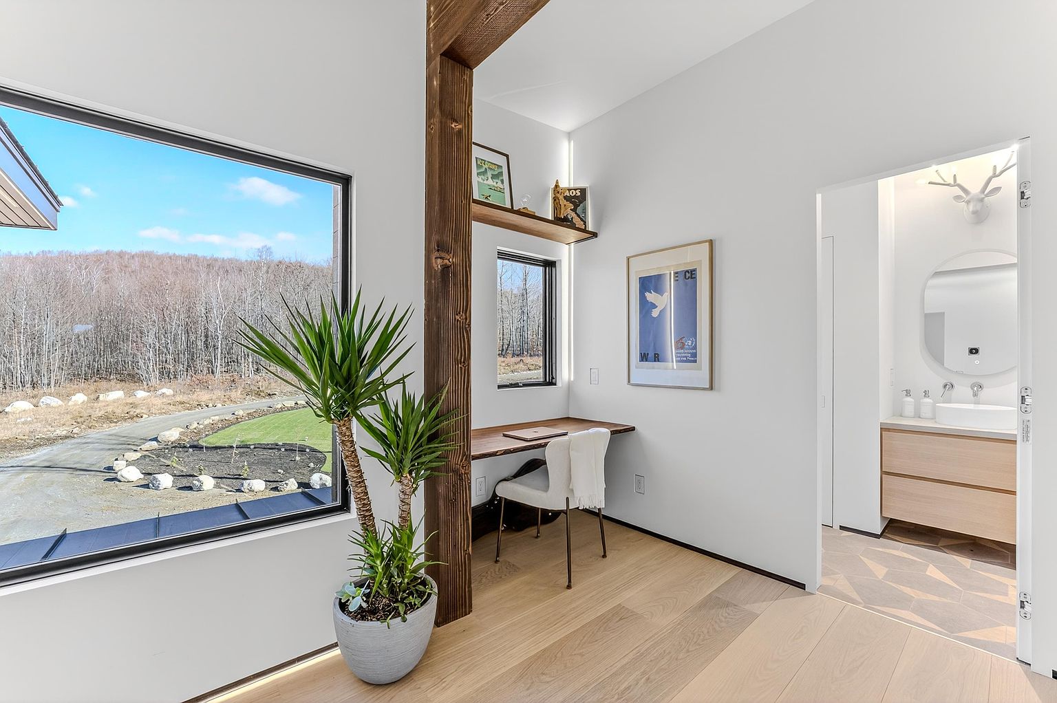 This bright and modern home office nook features a built-in wooden desk set against a large window with scenic forest views. The space is accented by a rustic wooden support beam, a potted plant, and minimalist decor, creating a serene and productive atmosphere. A doorway leads into a clean, contemporary bathroom, highlighting the home's cohesive and stylish design.
