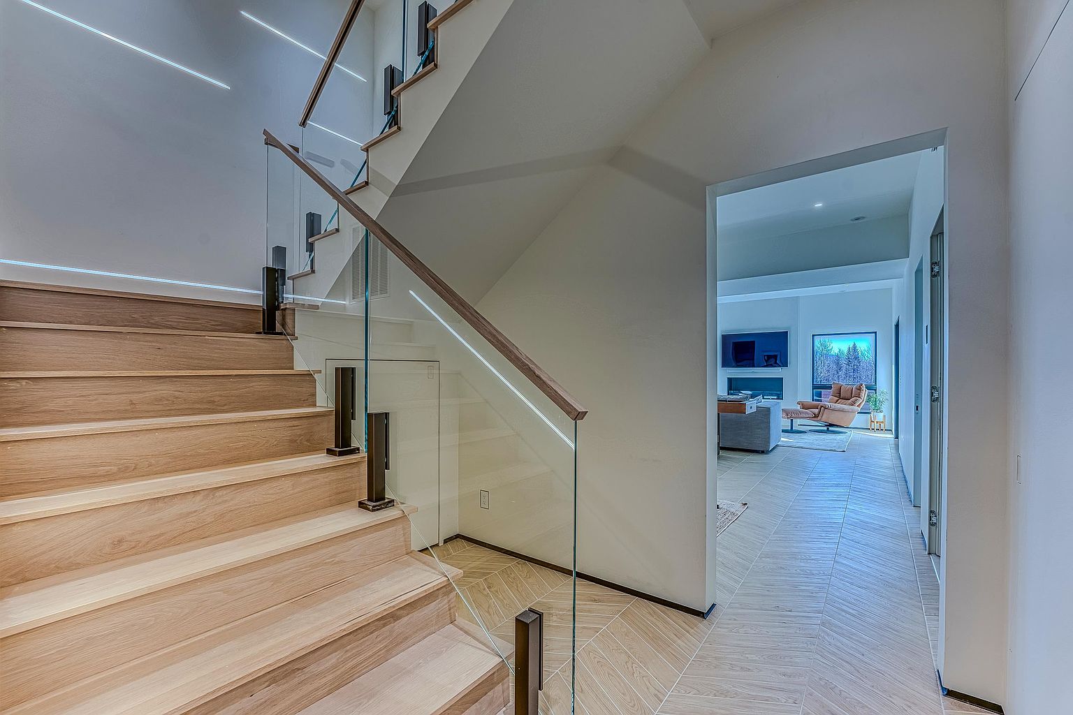 This image captures a modern, minimalist staircase featuring light wood treads and a sleek glass railing with dark metal accents. The perspective looks down the stairs toward a bright, open-concept living area with herringbone-patterned tile flooring, creating a seamless transition between levels. The clean lines and neutral color palette emphasize a contemporary architectural style.