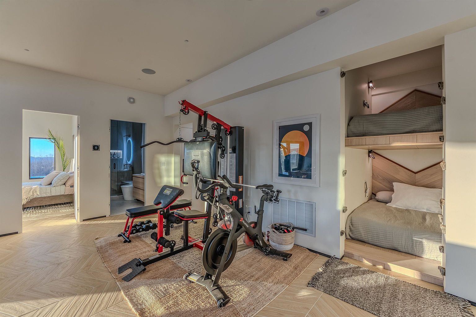 This versatile home gym space features a multi-functional workout station and a Peloton bike set on a textured area rug. To the right, a built-in bunk bed nook with warm wood paneling adds a unique, space-saving design element, while an open doorway leads to a bright, adjacent bedroom and bathroom. The room is finished with light-toned herringbone flooring and modern recessed lighting, creating a clean and functional multi-purpose environment.