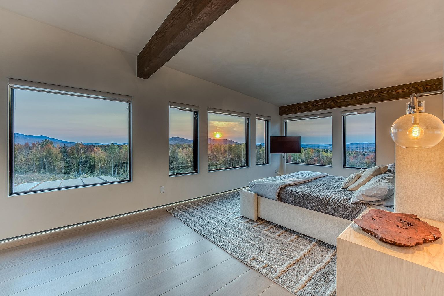 This serene primary bedroom features a minimalist design with a low-profile bed and a textured area rug, emphasizing a calm and sophisticated atmosphere. Large, expansive windows frame a breathtaking panoramic view of a forested landscape at sunset, seamlessly blending the interior with the natural surroundings. The room is accented by warm wooden ceiling beams and a unique, organic-shaped wooden decorative piece on a light-toned nightstand, creating a luxurious and tranquil retreat.