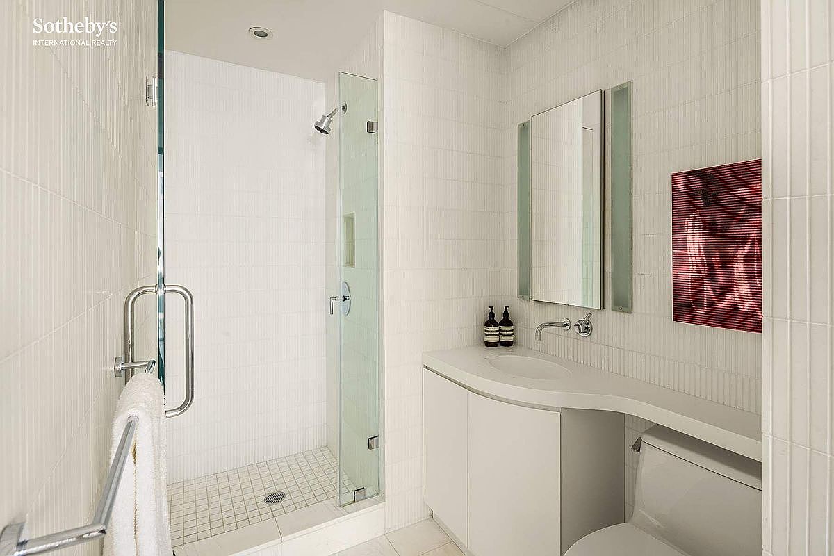 This modern, compact bathroom features a sleek, curved vanity with a white countertop and integrated sink, paired with a frameless glass-enclosed shower. The walls are finished with clean, vertical-patterned white tiles, creating a bright and minimalist aesthetic. A rectangular mirror hangs above the vanity, complemented by a piece of abstract red art that adds a bold pop of color to the otherwise neutral space.