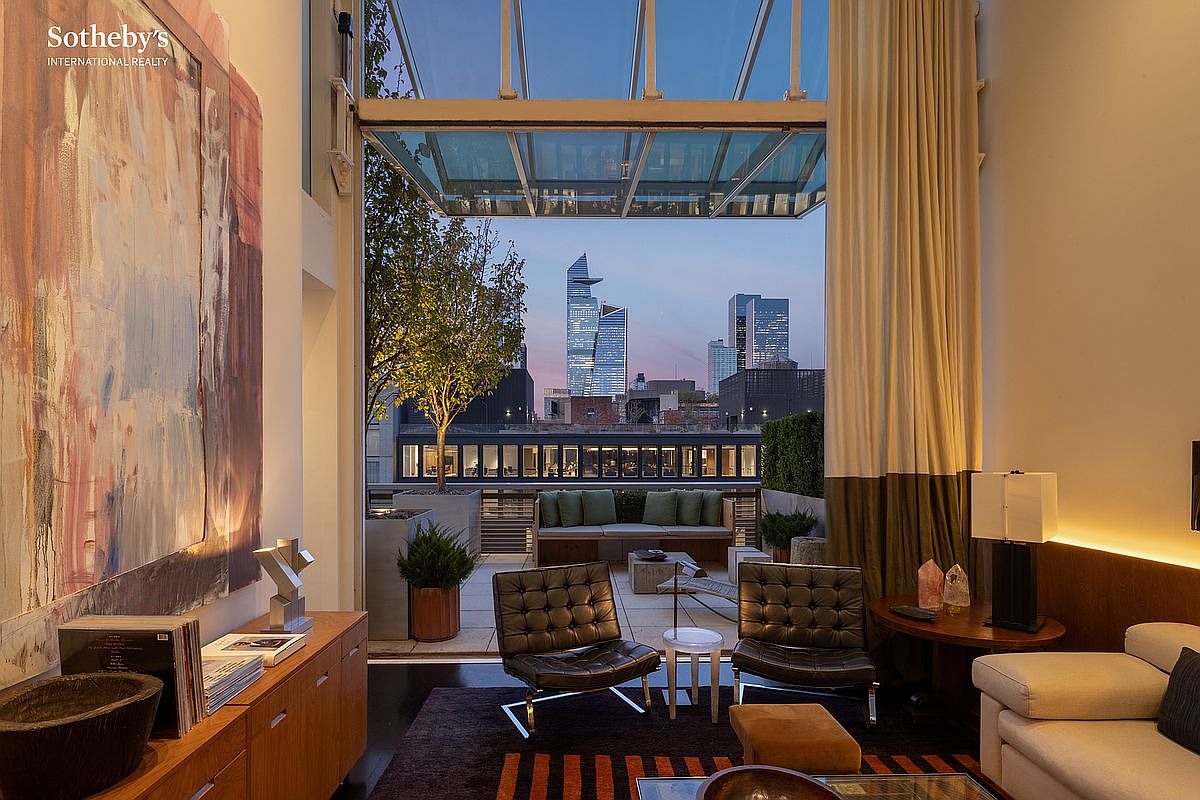 This sophisticated living room seamlessly transitions into an expansive outdoor terrace through a massive, retractable glass wall, offering a stunning panoramic view of the city skyline at dusk. The interior features high-end mid-century modern furniture, including iconic leather lounge chairs and a sleek wooden console, complemented by a large, vibrant abstract painting. The space exudes a luxurious, urban atmosphere, perfectly blending indoor comfort with an impressive outdoor entertainment area.