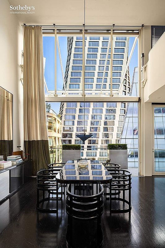 This sophisticated dining room features a sleek, dark glass-topped table surrounded by modern, curved black chairs, set against a dramatic floor-to-ceiling window wall. The expansive glass provides a striking urban view of a neighboring high-rise, while a tall, two-toned curtain adds a touch of elegance to the double-height space. The dark hardwood floors and minimalist aesthetic create a luxurious, contemporary atmosphere perfect for high-end city living.