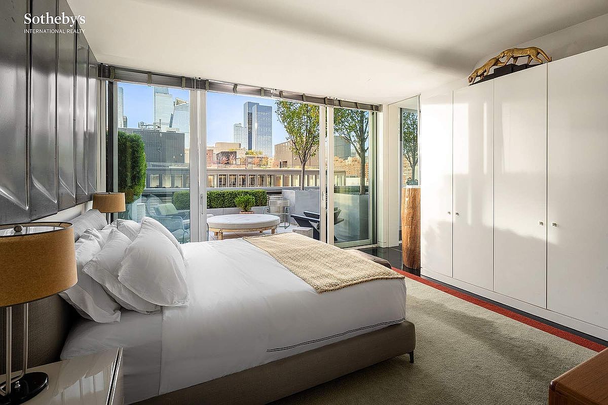 This sophisticated primary bedroom features a plush king-sized bed with crisp white linens and a neutral-toned throw, positioned to face floor-to-ceiling glass sliding doors that open onto a private terrace. The room is accented by a sleek, white floor-to-ceiling wardrobe wall and a modern gold-toned sculpture perched atop it, creating a clean, minimalist aesthetic. The perspective captures the seamless transition between the luxurious interior and the urban cityscape visible through the expansive windows.