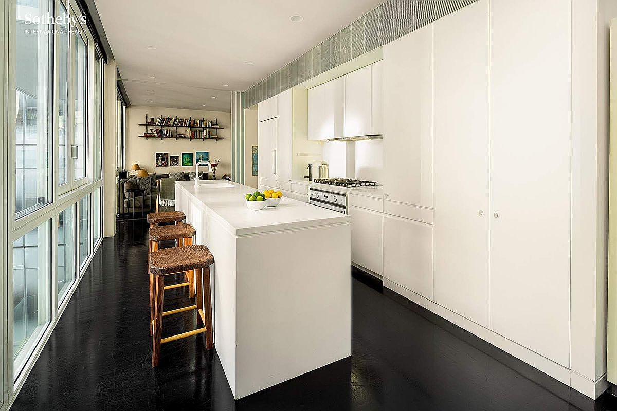 This sleek, modern kitchen features a minimalist design with crisp white cabinetry, a large central island with a white countertop, and dark, polished flooring that creates a striking contrast. The space is illuminated by recessed lighting and natural light streaming through a long wall of floor-to-ceiling windows, which also provide a view into an adjacent living area. The overall aesthetic is clean, sophisticated, and highly functional, perfect for contemporary urban living.
