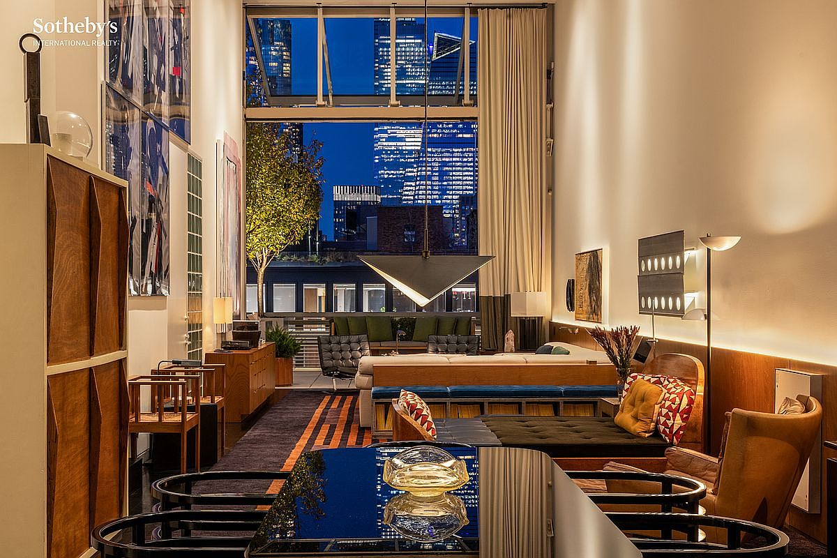 This sophisticated, double-height living space features floor-to-ceiling windows that frame a stunning urban skyline at dusk. The room is furnished with a mix of mid-century modern pieces, including a plush sofa, a low-profile daybed, and a sleek dining table in the foreground, all set against warm wood paneling and high-end art. The perspective is a wide, eye-level shot that captures the seamless transition between the luxurious interior and the vibrant city view outside.