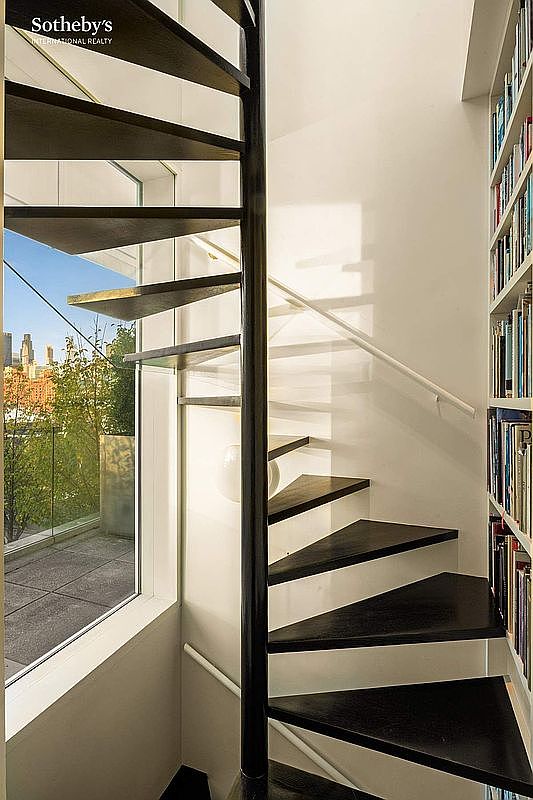 This image captures a sleek, modern spiral staircase with dark wood treads and a central black metal pole, set against a crisp white wall. To the right, a floor-to-ceiling built-in bookshelf adds a sophisticated touch, while a large window to the left provides a bright, airy view of an outdoor terrace and city skyline. The composition emphasizes clean lines and a minimalist aesthetic, creating a striking architectural focal point.