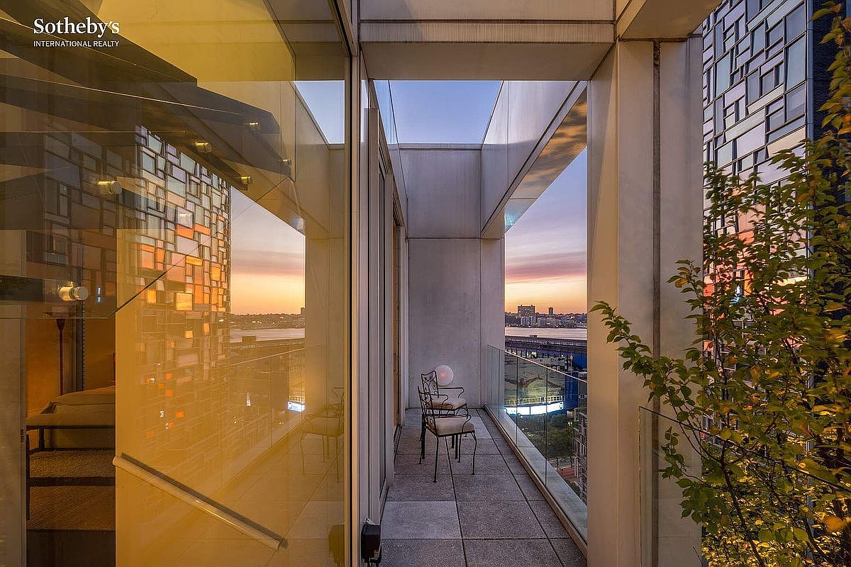 This sleek, modern balcony offers a sophisticated outdoor living space with a glass railing that maximizes the panoramic city and waterfront views. The area is furnished with minimalist metal chairs and features a small potted tree, creating an intimate setting perfect for enjoying the sunset. The architectural design incorporates clean lines and expansive glass walls, seamlessly blending the indoor and outdoor environments.