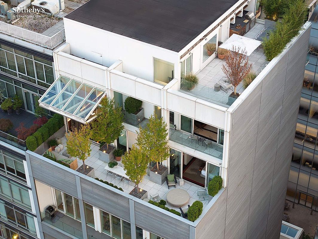 This high-angle aerial view captures a sophisticated multi-level urban penthouse terrace, showcasing a seamless blend of modern architecture and lush landscaping. The expansive outdoor space features multiple seating areas, dining arrangements, and mature trees in sleek planters, all set against a backdrop of contemporary glass-and-steel building design. The perspective emphasizes the luxury and exclusivity of this private outdoor oasis in a dense city environment.
