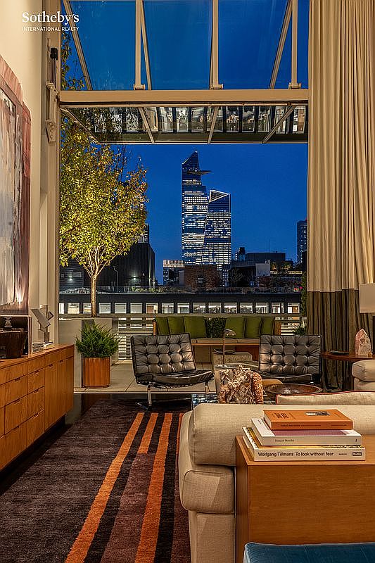 This sophisticated living room features a seamless transition to an outdoor terrace through a massive, retractable glass wall that frames a stunning nighttime view of the New York City skyline. The interior is elegantly furnished with mid-century modern leather lounge chairs, a plush sofa, and a rich, textured area rug with bold orange stripes. The space exudes a luxurious, urban atmosphere, perfectly blending high-end indoor comfort with an iconic metropolitan backdrop.